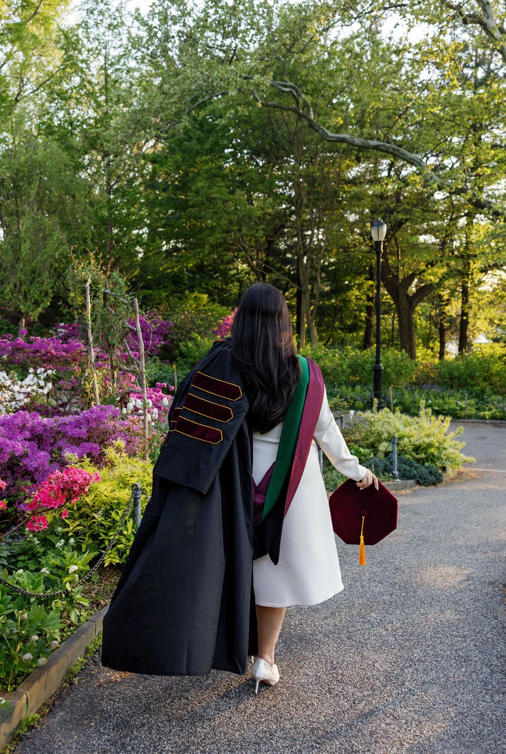 Graduate in black doctoral gown with maroon hood walks through lush garden path holding graduation cap.