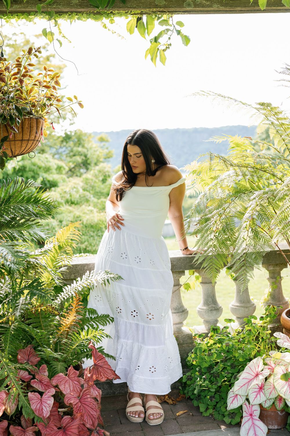 White eyelet dress and sandals styled in a lush garden setting with pink flowers and stone balustrade details.