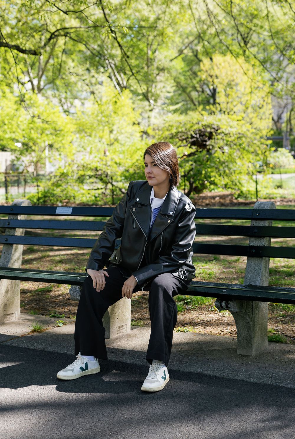 Person in black leather jacket and pants sitting on a park bench surrounded by lush green trees.