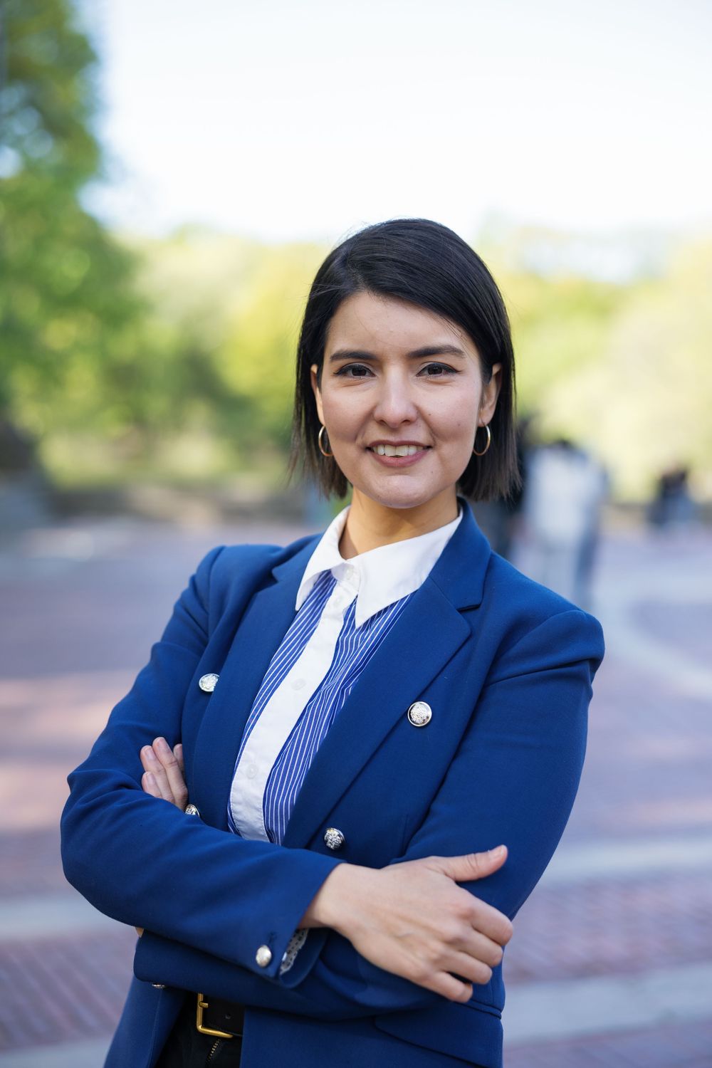 Professional woman in blue blazer with arms crossed smiling outdoors on a brick pathway.