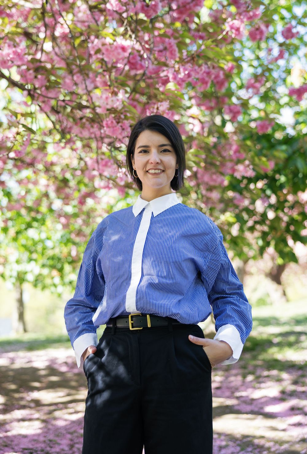 Woman in blue striped shirt smiling outdoors under pink cherry blossom trees in spring.