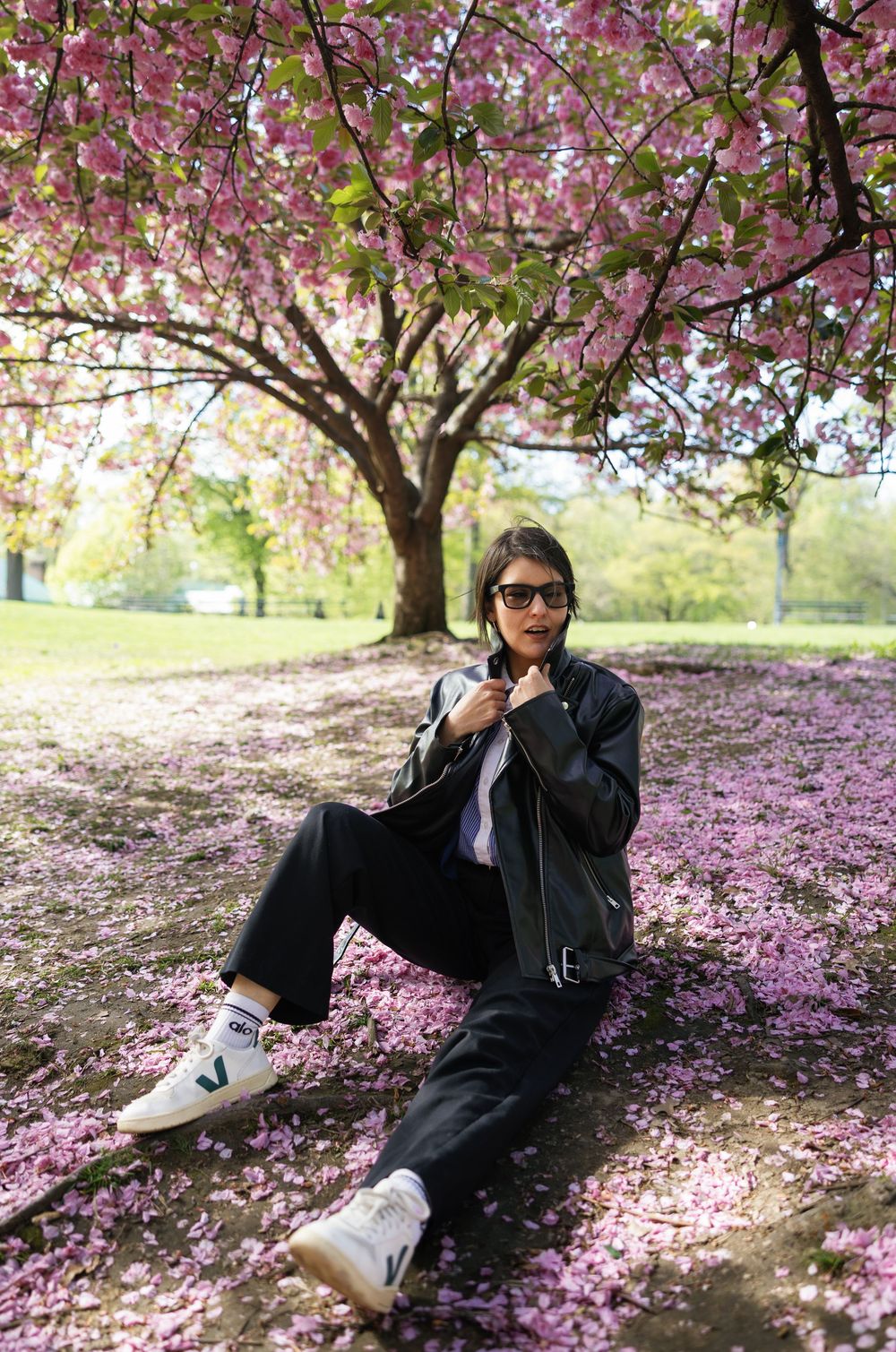 Person sitting under a blooming pink cherry blossom tree surrounded by fallen petals wearing a black leather jacket and white sneakers.