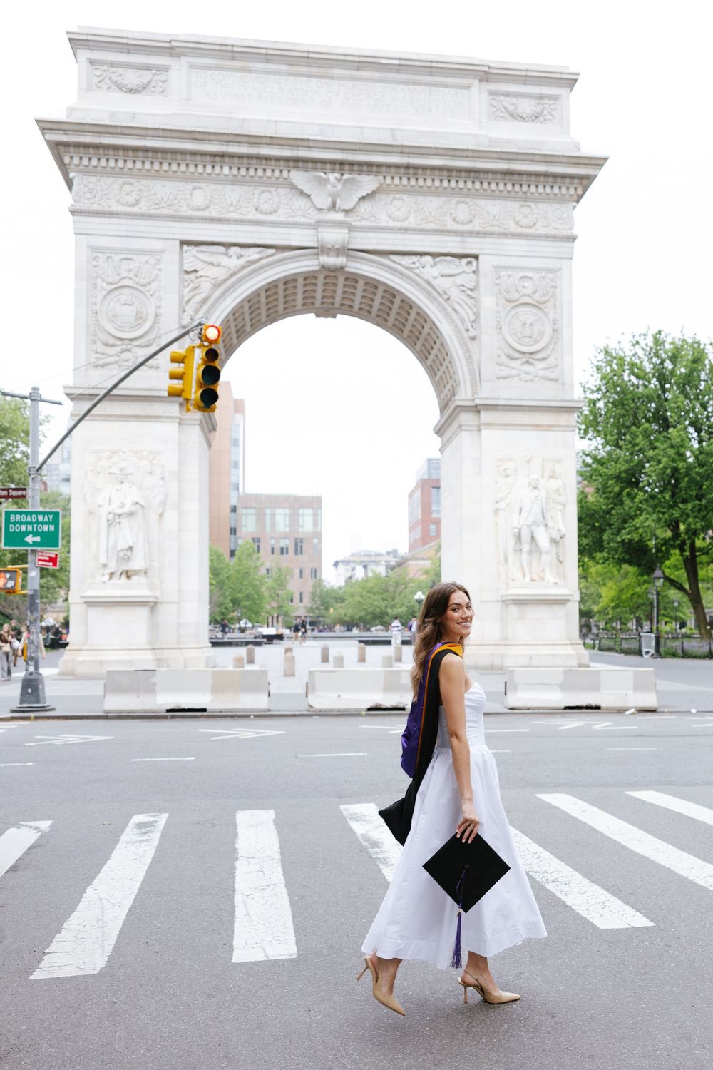 Graduate in white dress holding cap walks at Washington Square Park arch in New York City.