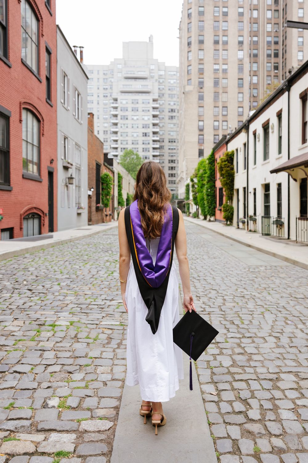 Graduate in white dress and purple sash walks cobblestone street holding mortarboard cap in urban setting.