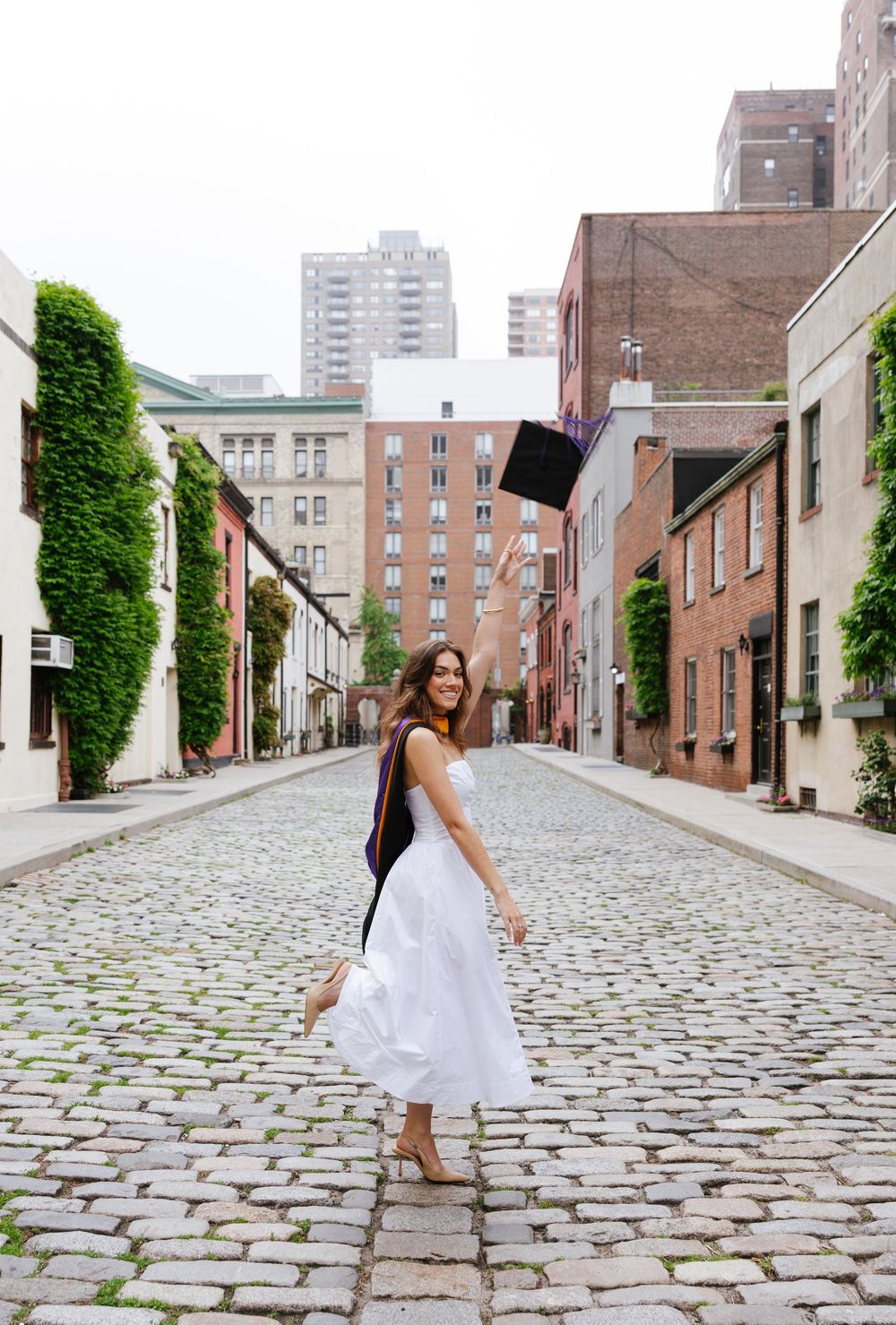 A graduate in a white dress tosses her cap on a cobblestone street lined with brick buildings and ivy.