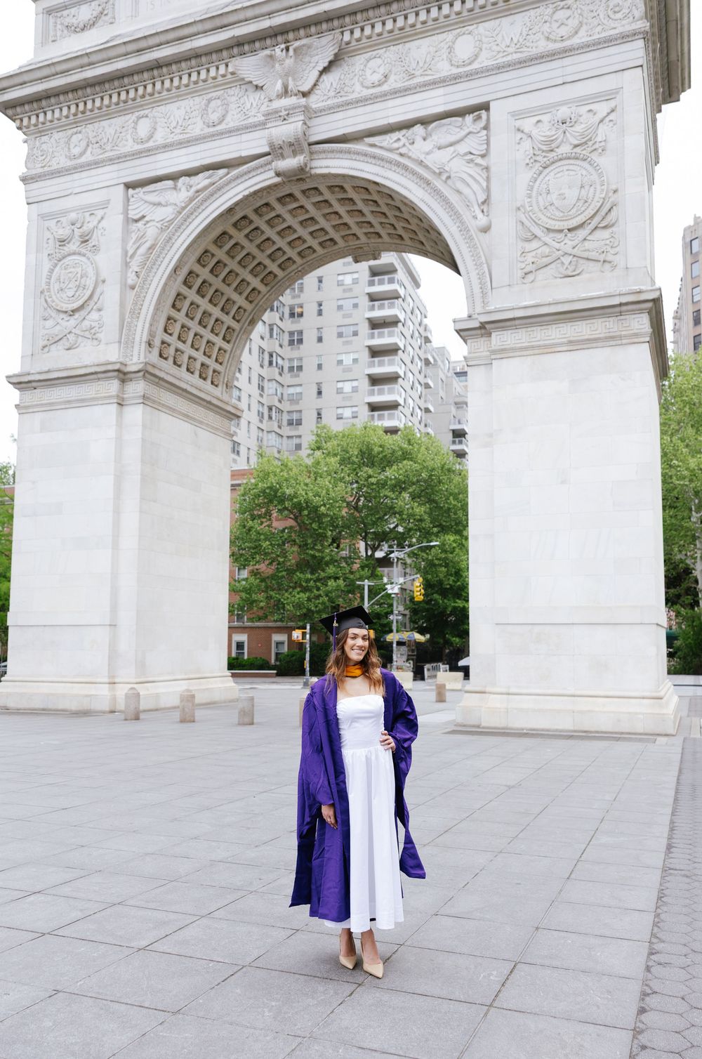 Graduate in purple cap and gown poses before Washington Square Arch in NYC.