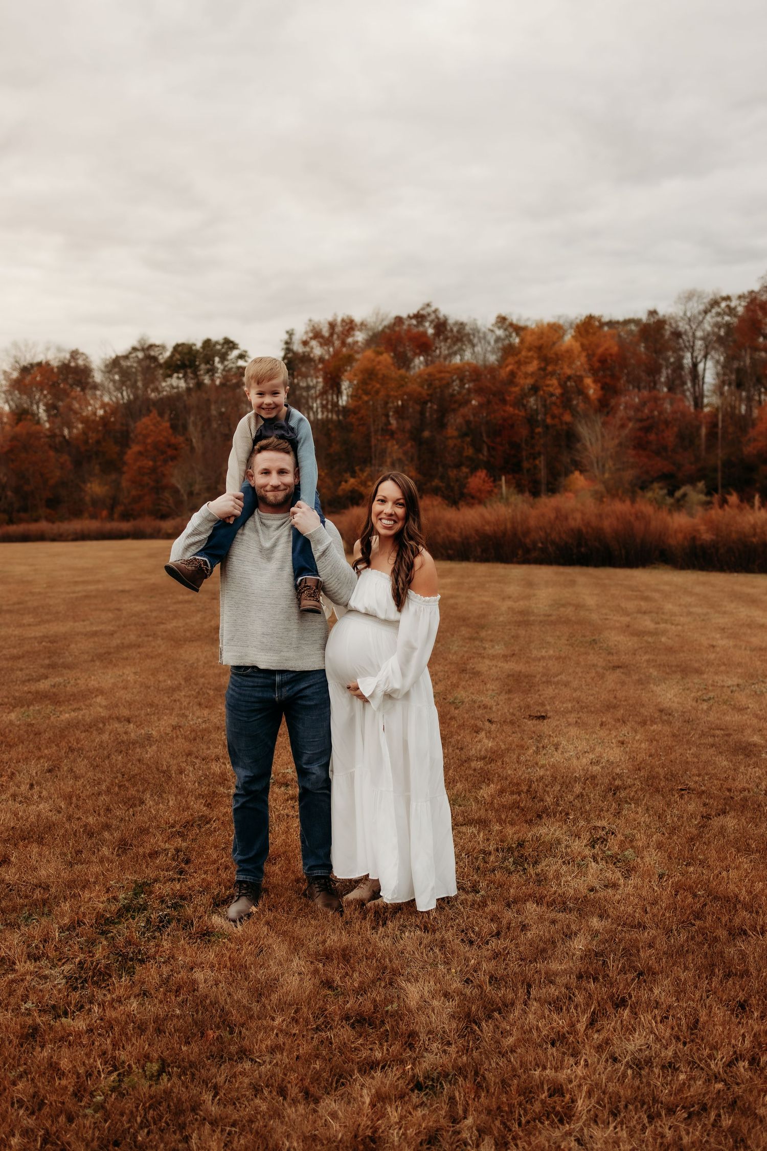 A couple stands together in a rustic autumn field with orange foliage in the background.