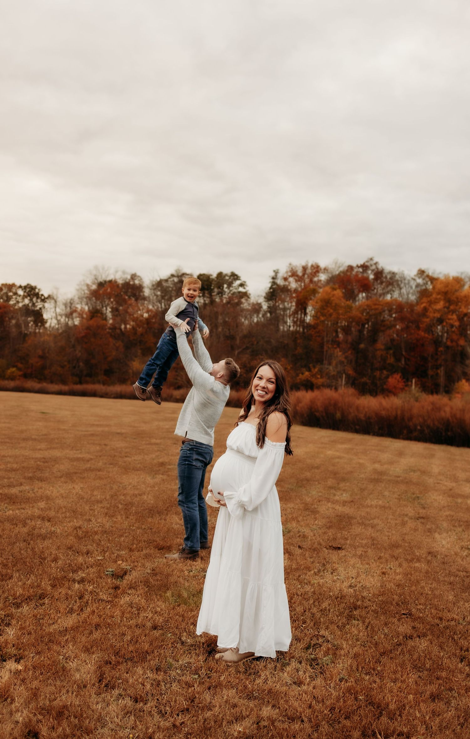 A couple in a white dress and casual attire dances together in an autumn field with orange trees in the background.