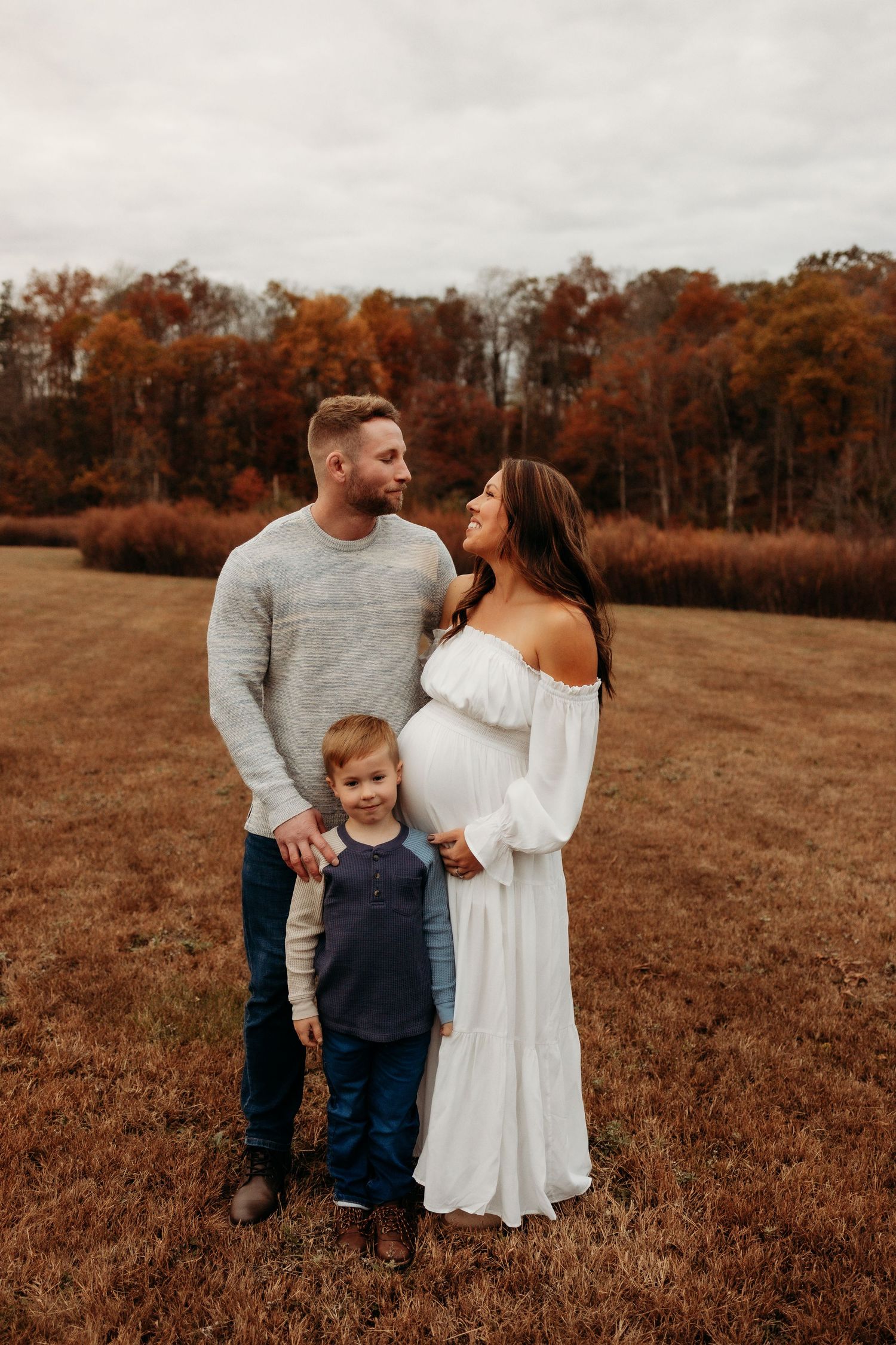 Family in autumn field wearing white and grey casual outfits share candid moments during outdoor photo session.