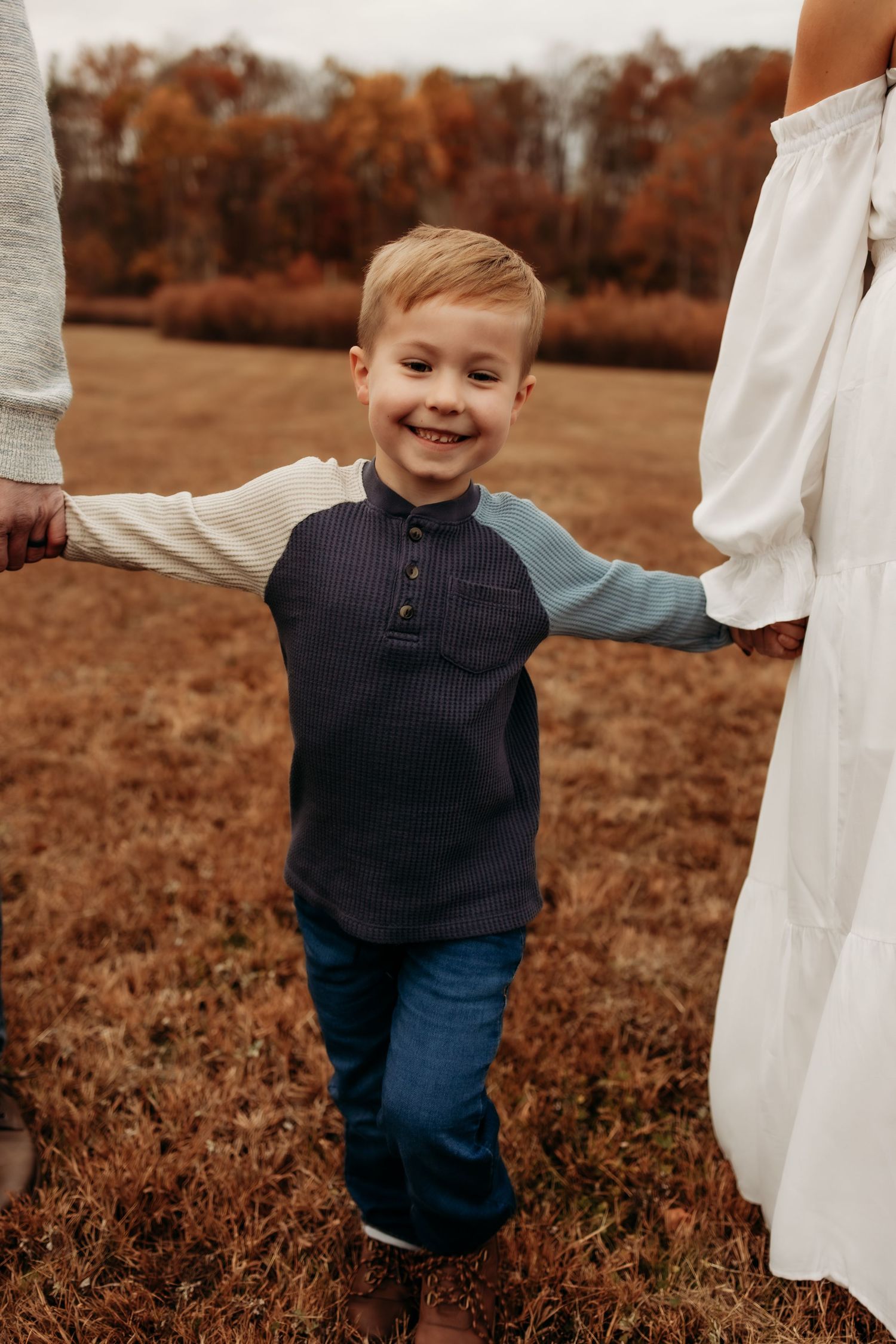A child wearing a navy henley shirt and jeans holds hands with family members in an autumn field at sunset.