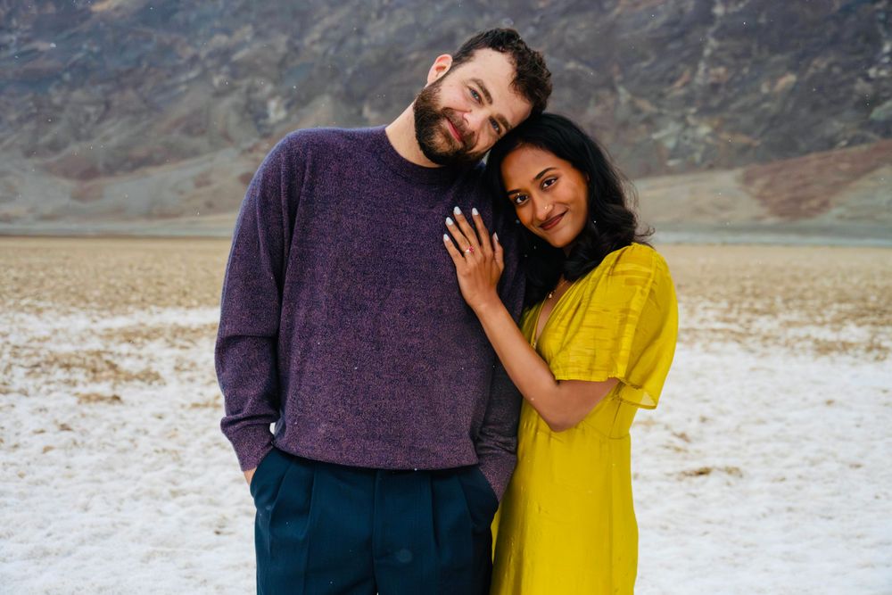Smiling couple embracing in a vast white salt flat with dramatic mountain backdrop behind them.