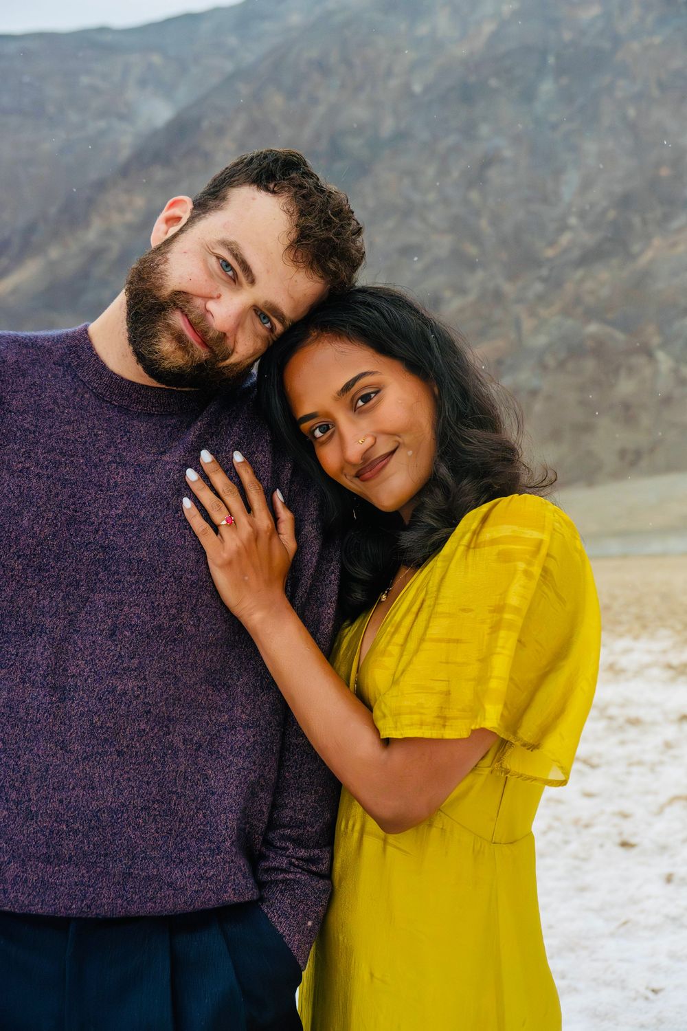 Woman in yellow dress leans on bearded man in purple sweater with hand on his chest outdoors.