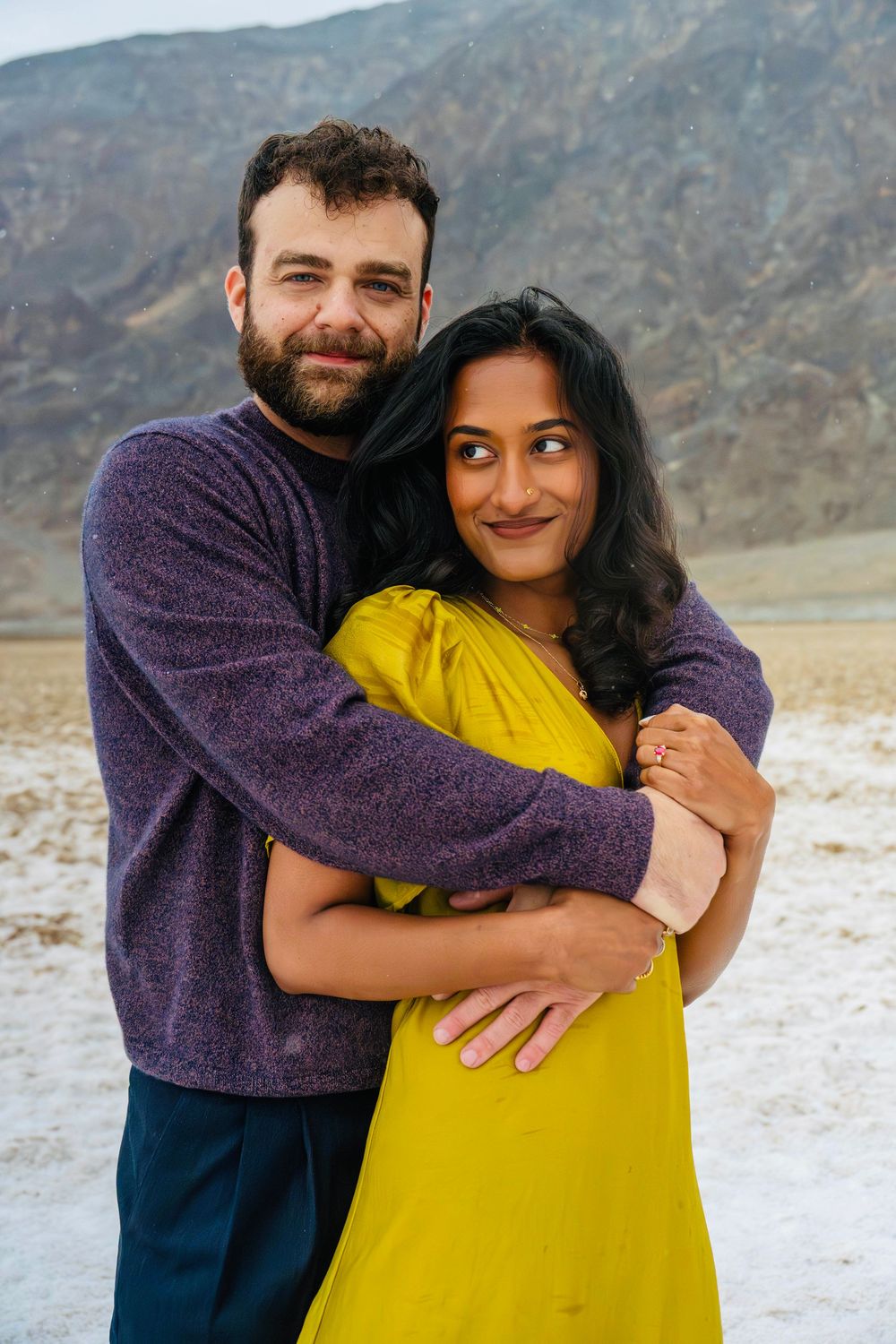 Bearded man hugging woman in yellow dress with mountains and white salt flats in the background.