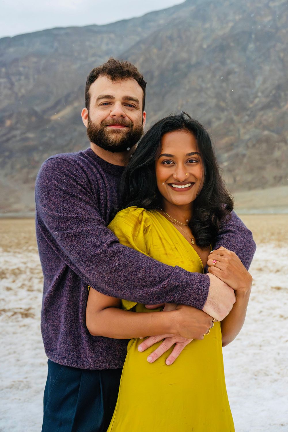 Couple embracing in Death Valley, man in purple sweater hugging woman in yellow dress with mountains behind.