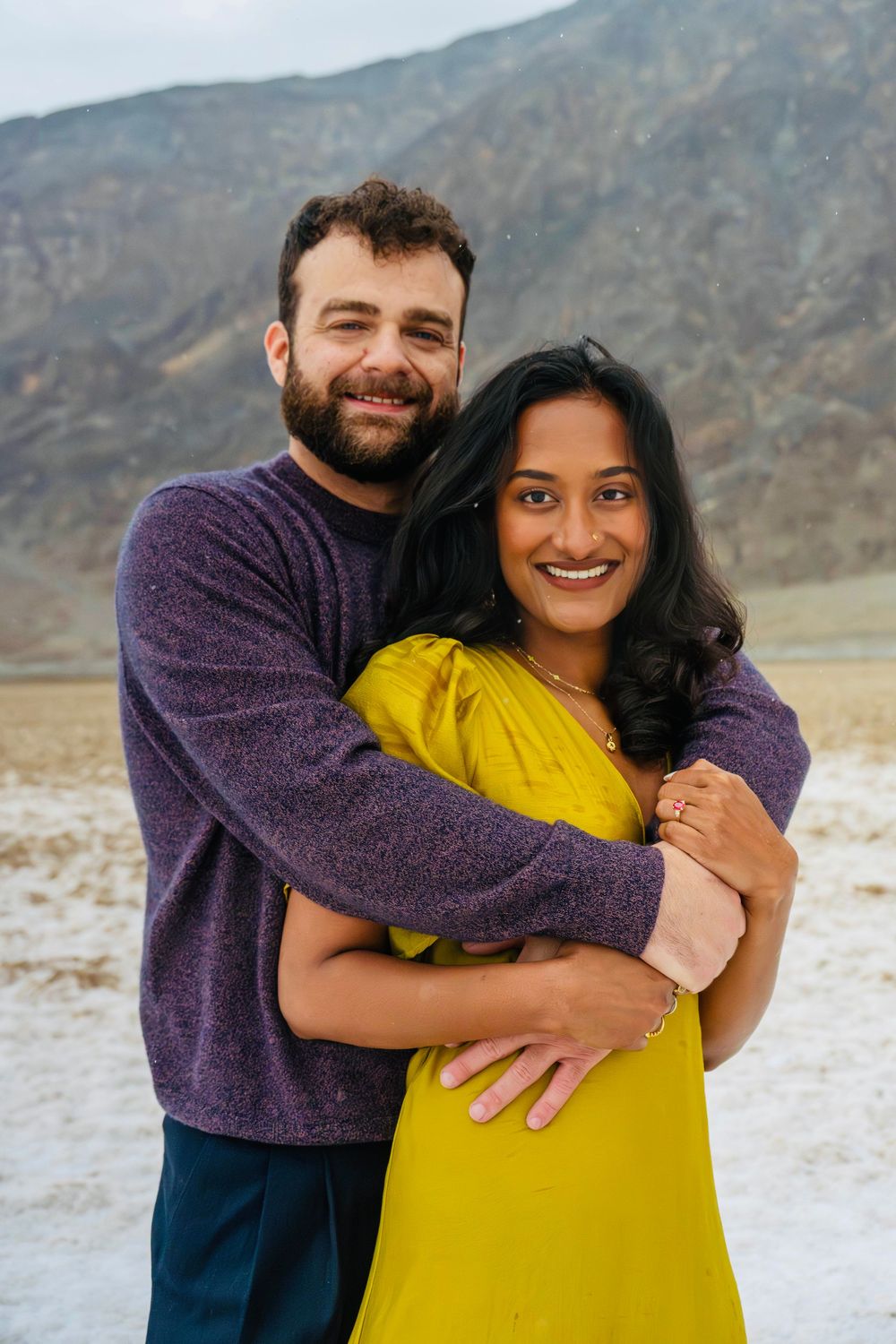 Happy couple posing together outdoors, man in purple sweater embracing smiling woman in yellow dress.