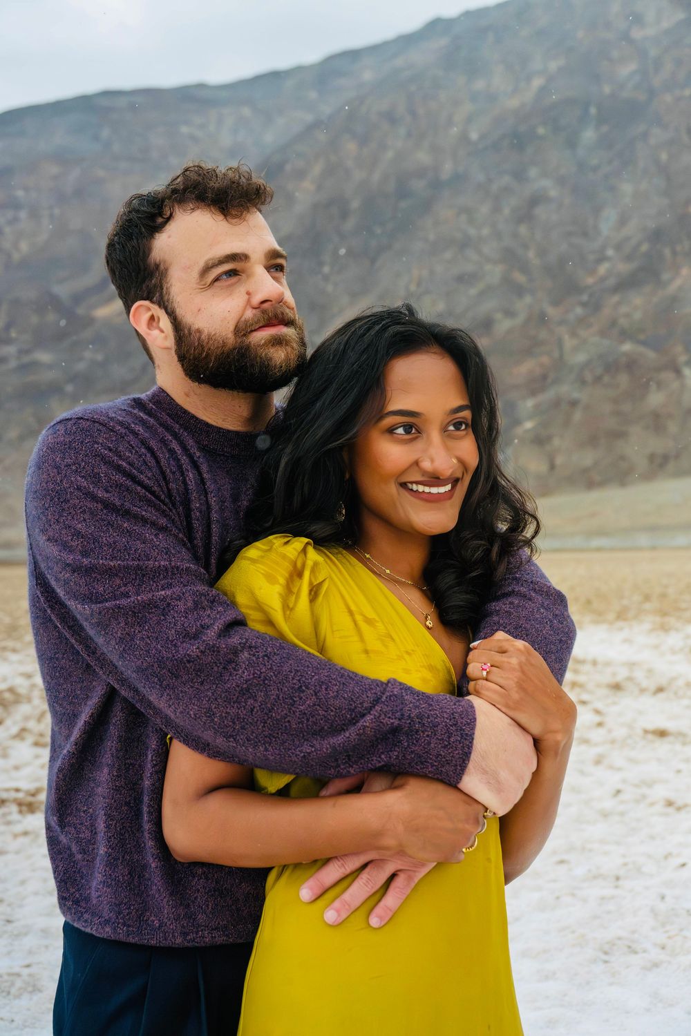 Smiling couple embracing in desert landscape, woman in yellow dress and man in purple sweater.