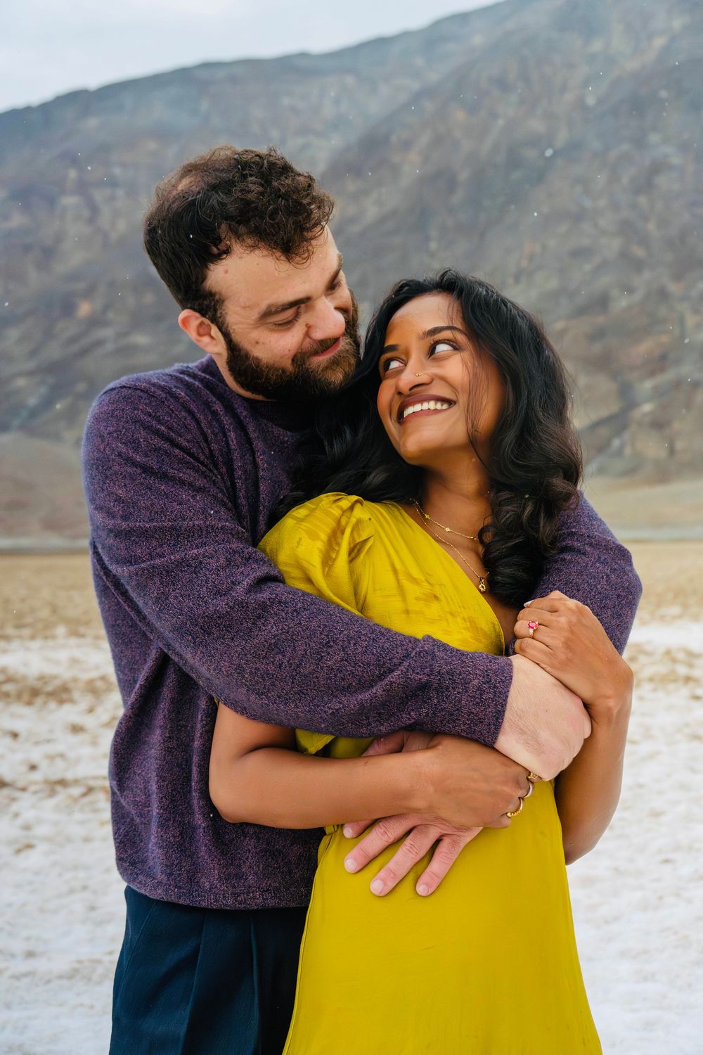 Couple sharing a joyful moment, woman in yellow dress laughing up at bearded man in purple sweater.