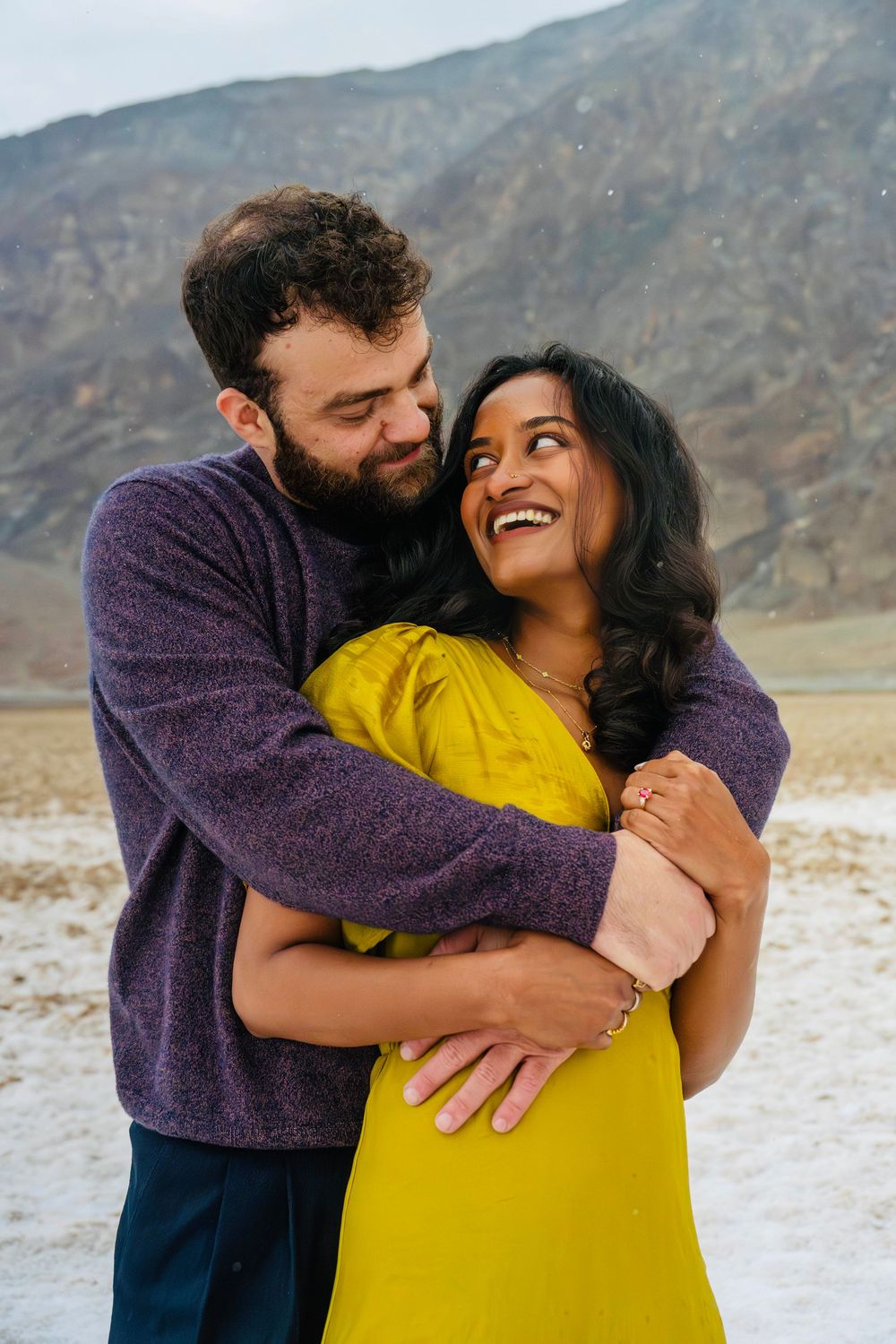 Laughing couple embracing outdoors in desert setting, woman in yellow dress and man in purple sweater.