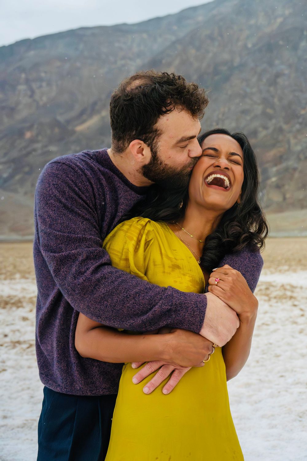 Man kissing woman on cheek as she laughs joyfully in desert landscape engagement photo.