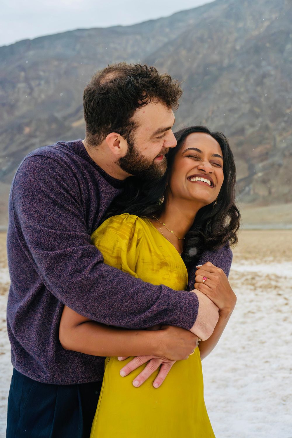 Couple sharing a romantic laugh outdoors in arid landscape, woman in yellow dress and man in purple.