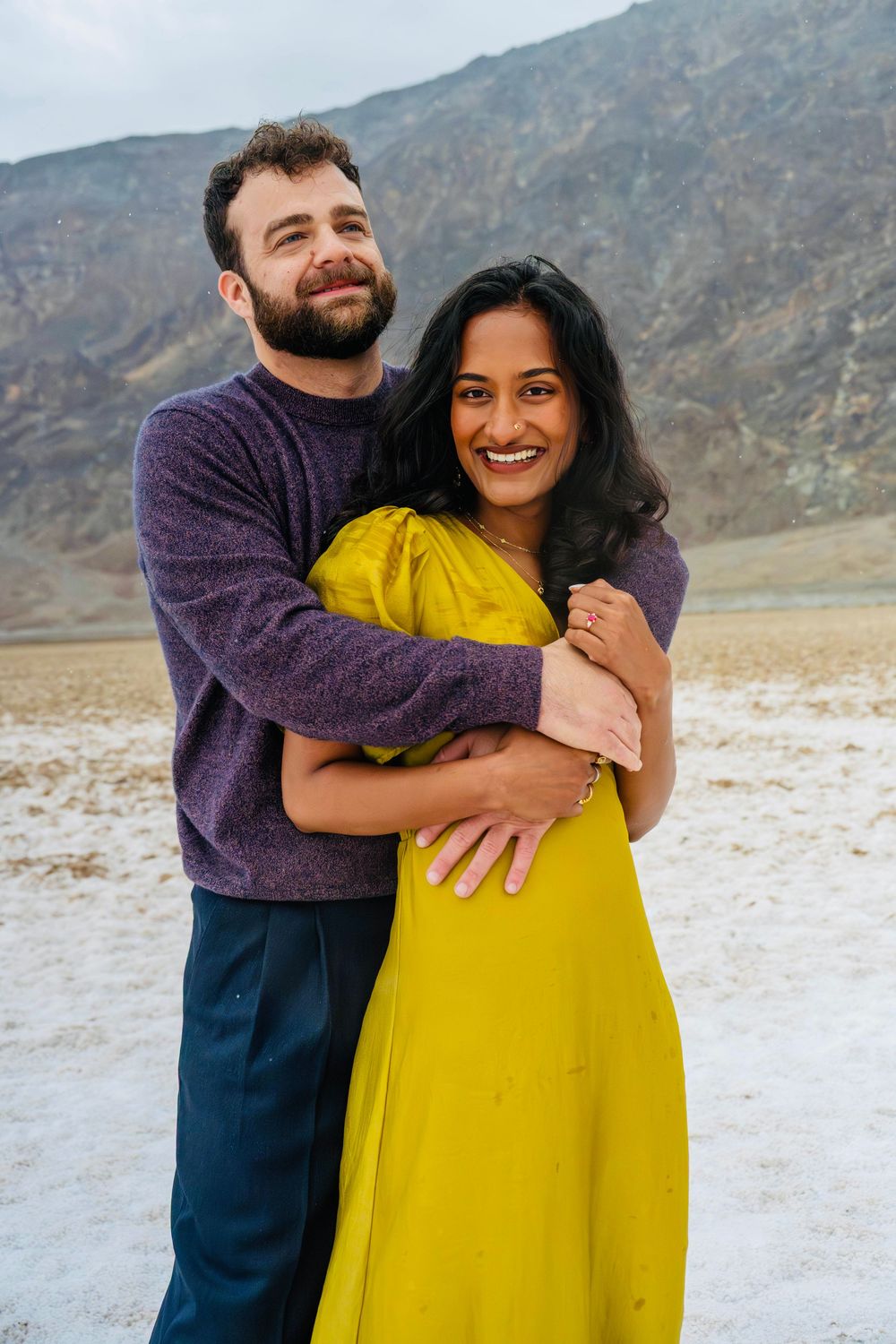 Happy couple posing in desert landscape, man in purple sweater hugging woman in bright yellow dress.