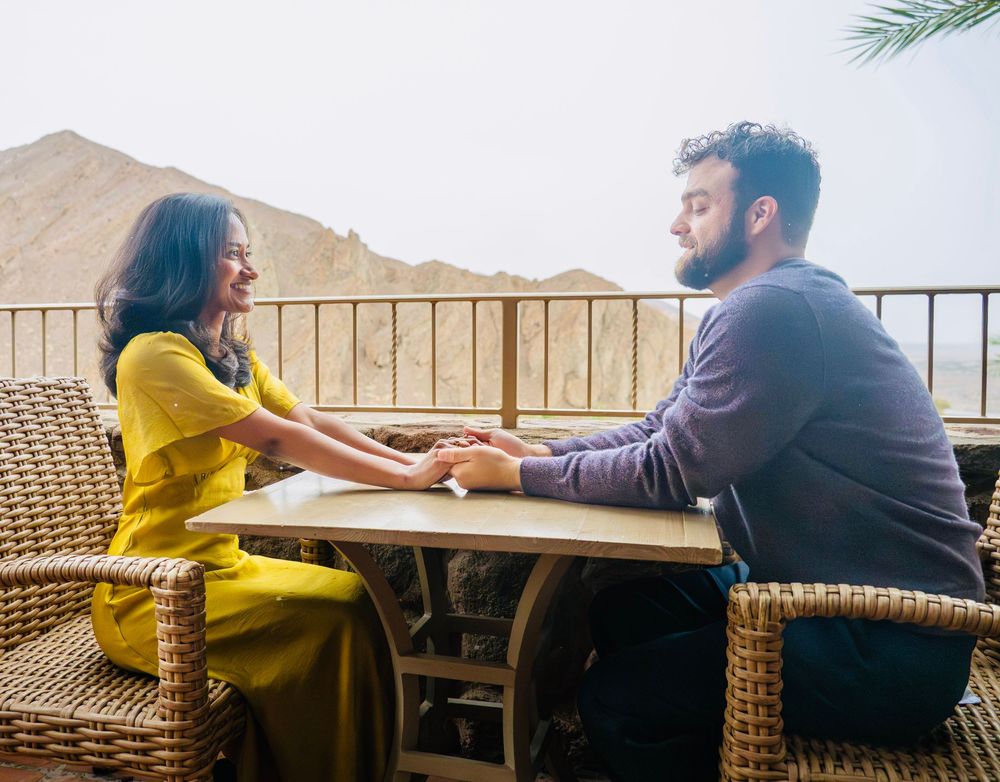 Couple holding hands across a wooden table on a scenic mountain balcony, woman in yellow dress smiling.