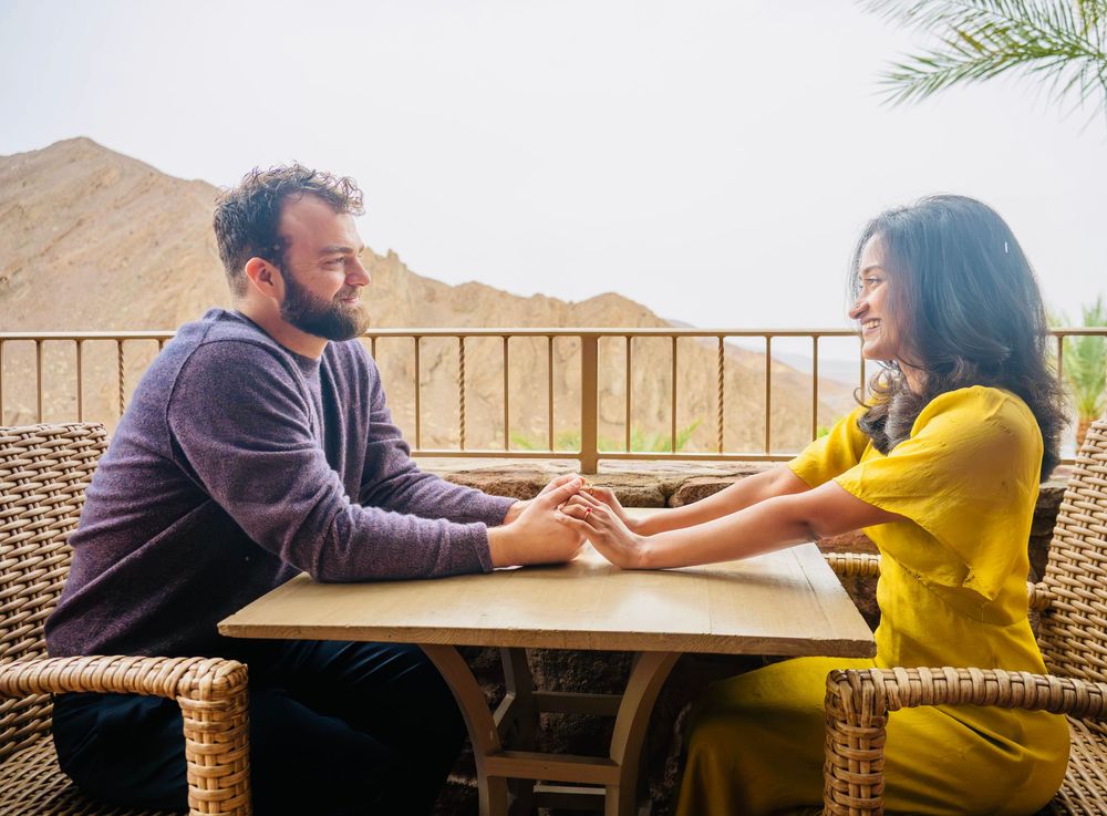 Romantic couple holding hands at outdoor table with mountain backdrop, man in purple sweater and woman in yellow.