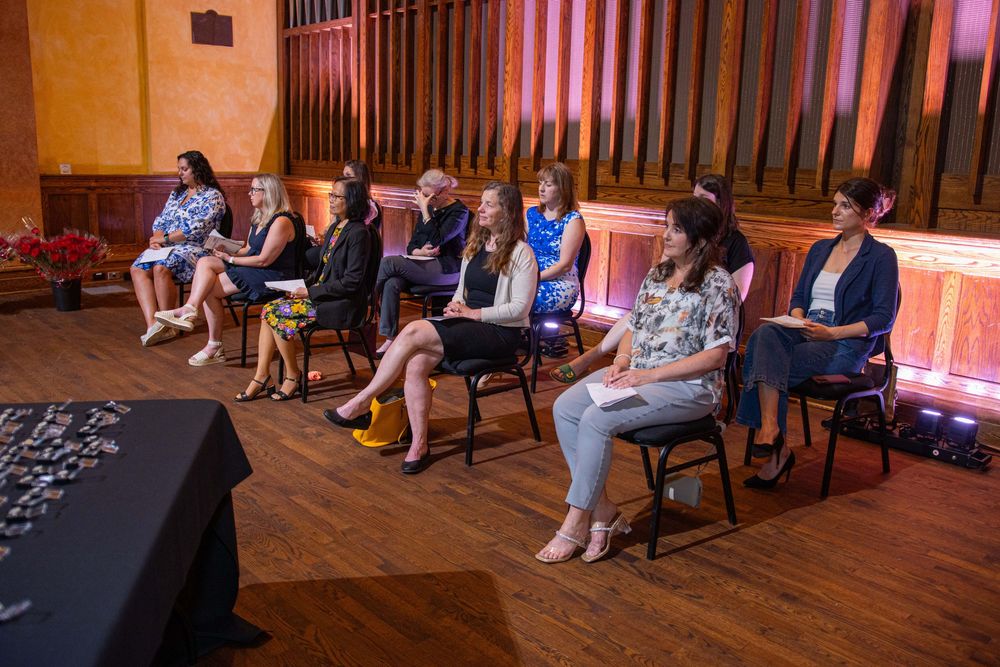 A row of people seated on chairs at an event with red-lit stage backdrop.
