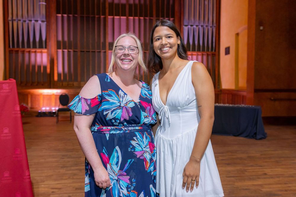 Two people in formal attire smile together at an indoor event with pink-lit organ pipes behind them.