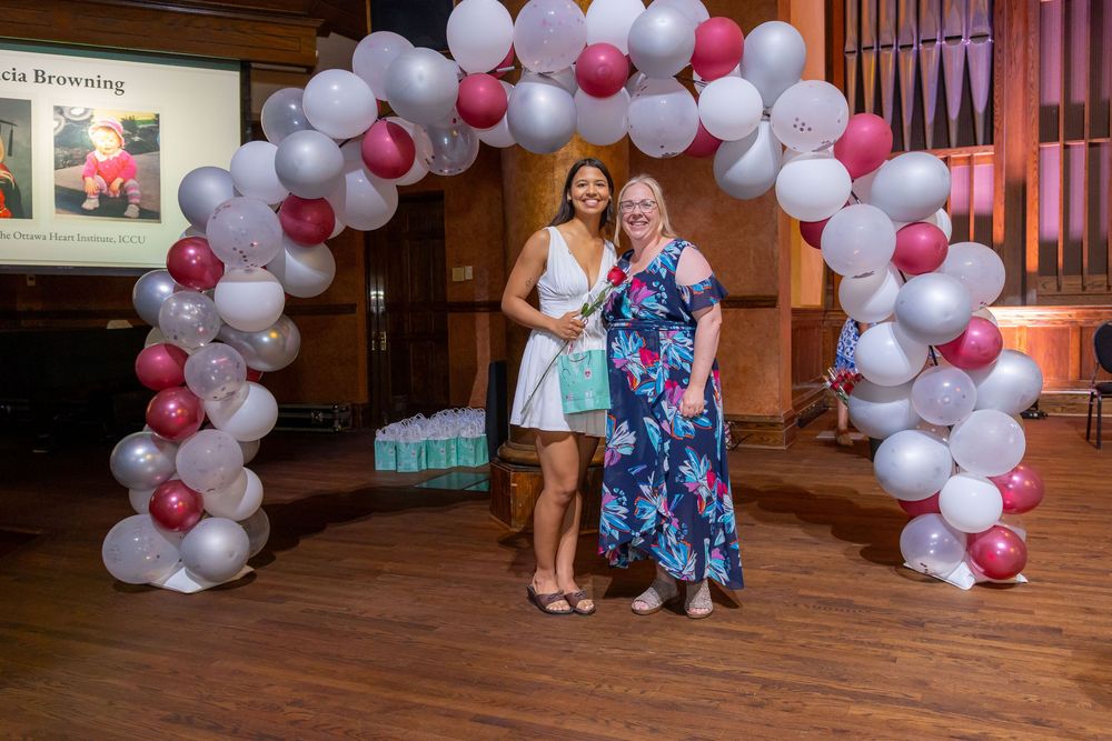 Two people pose under a pink and white balloon arch at a formal celebration.