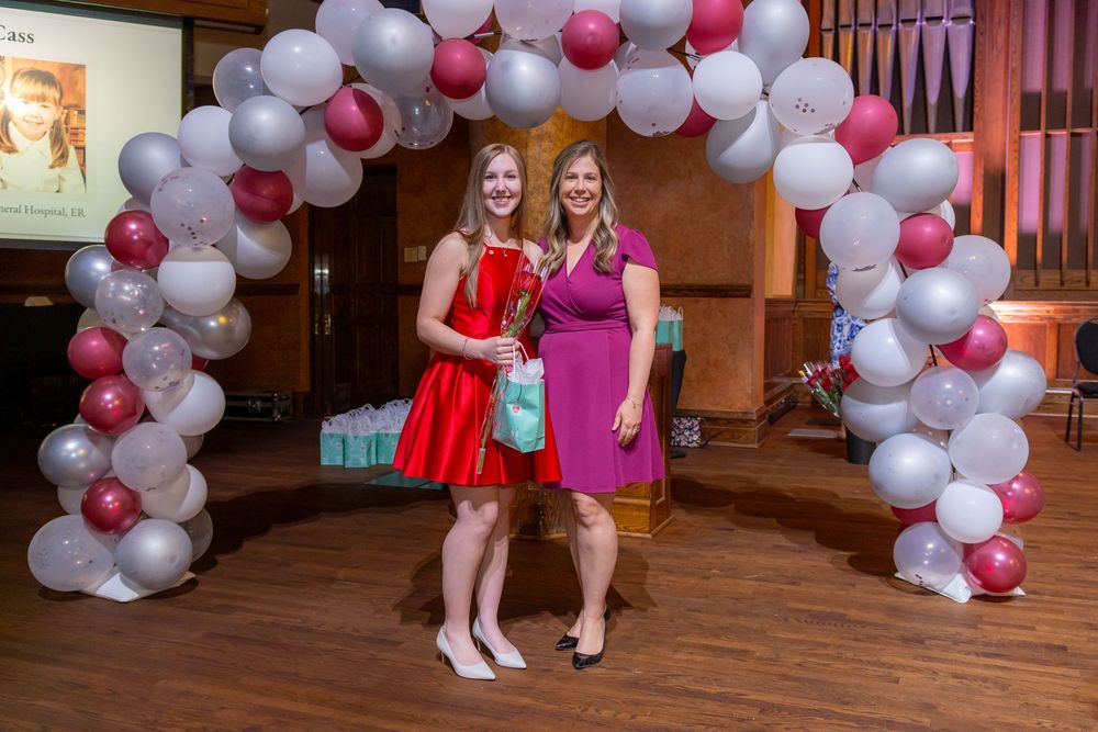 Two people in colorful dresses pose under a festive balloon arch at an indoor celebration.