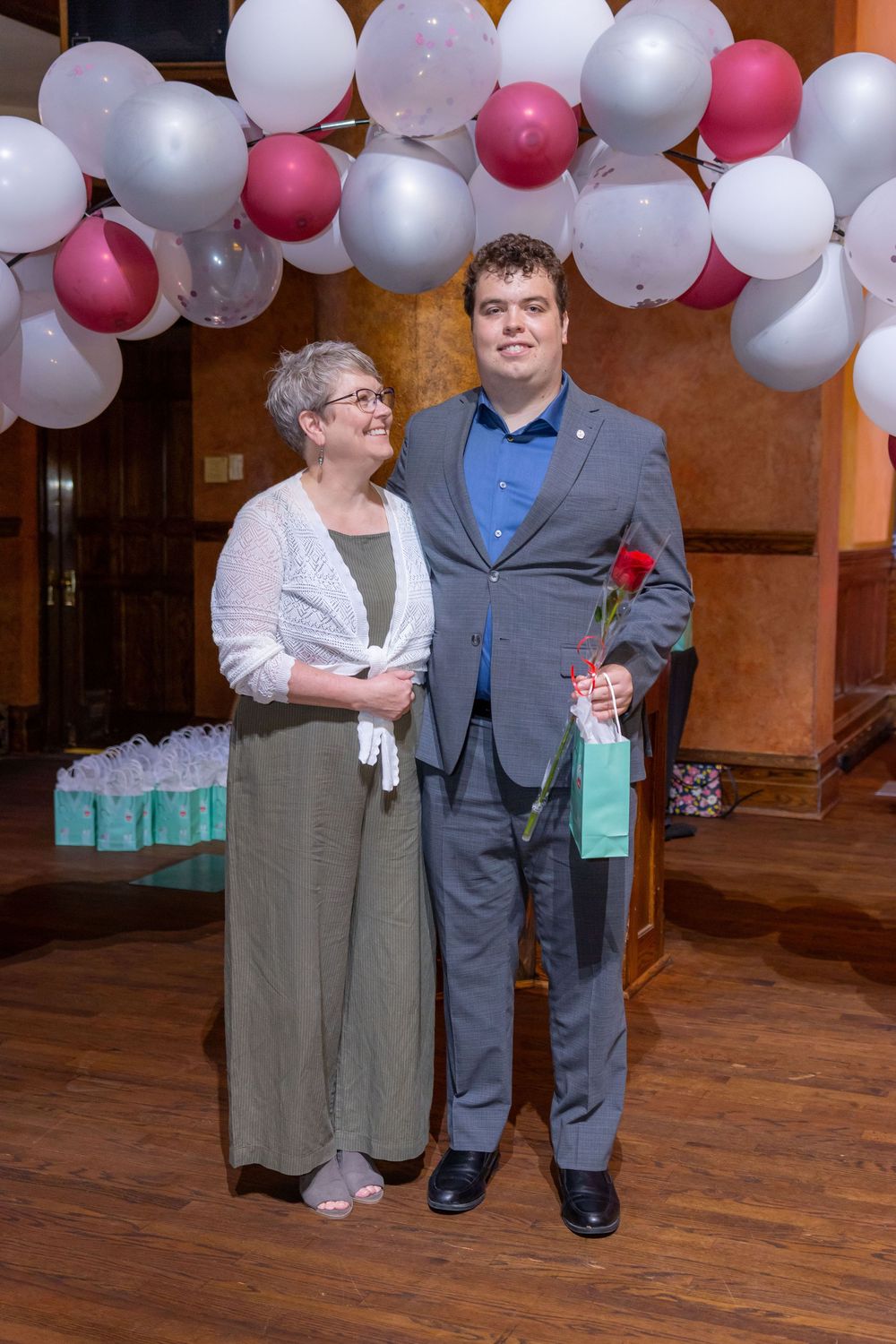 A couple poses under white and pink balloon arch at formal event with gifts visible.