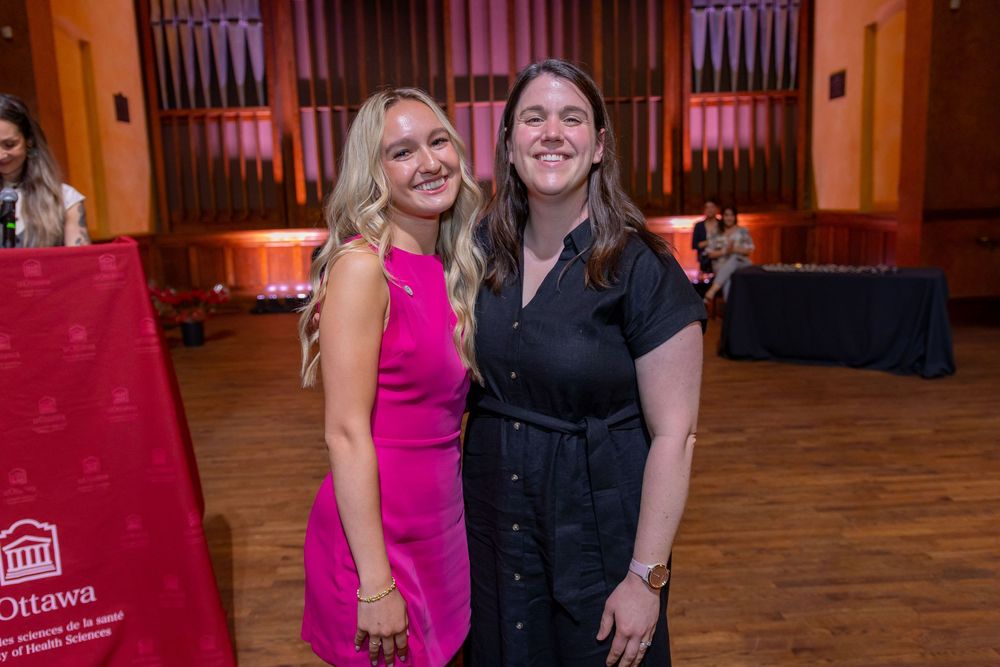 Two attendees pose together at an event, one wearing a bright pink dress and one in black.