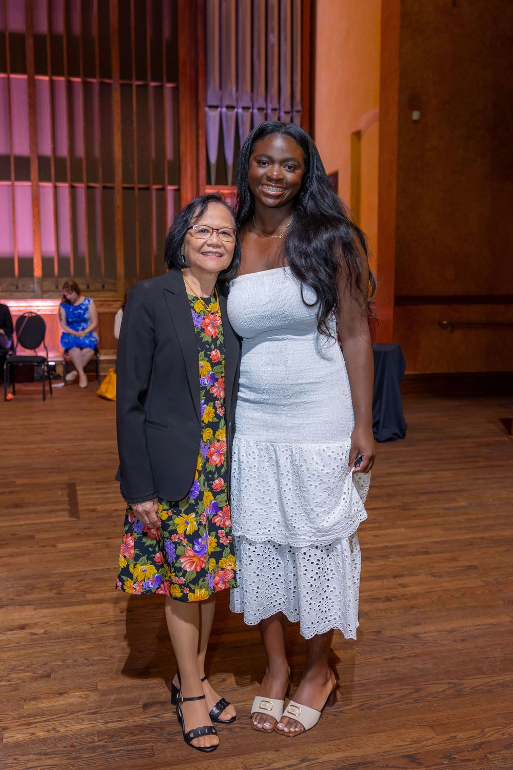 Two people pose together on a wooden floor at an event, one wearing a floral dress and the other in white.