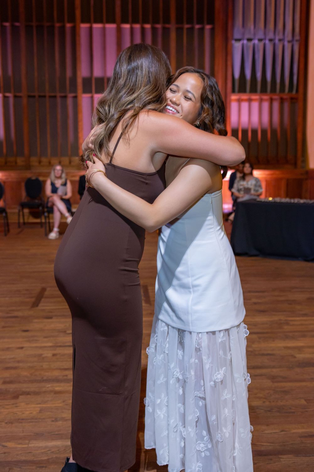 Two people share an emotional embrace on a wooden floor during what appears to be a special event.