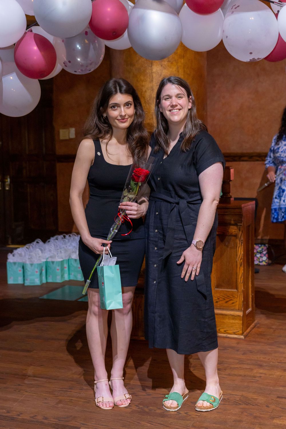 A pair of women stand close together on a hardwood floor with festive balloon decorations in the background.