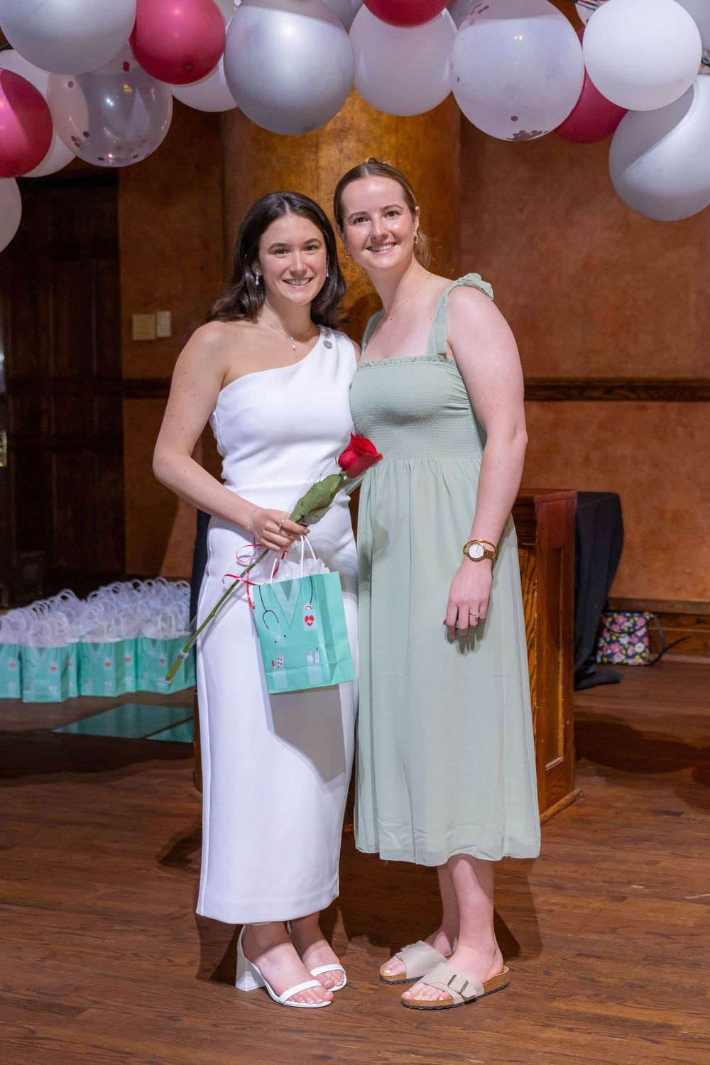 Two people in white and mint green dresses stand together on a wooden floor under red and white balloon decorations.