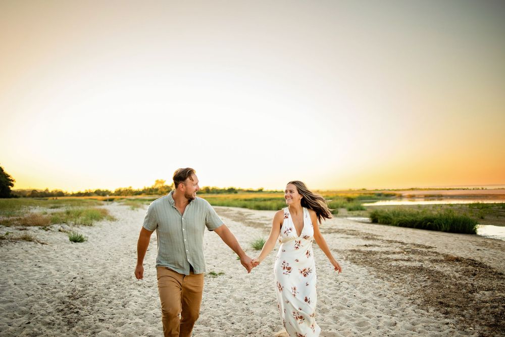 Couple walking hand in hand along a sandy beach at sunset with golden light illuminating the coastline.