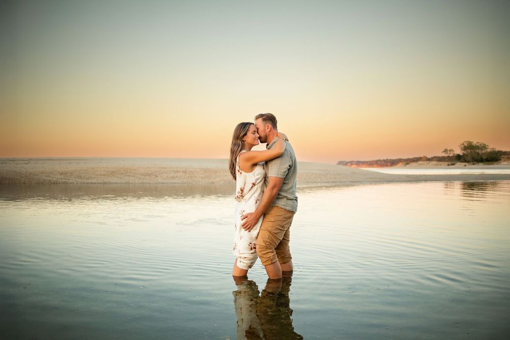 Two people embrace in the shallow ocean waters during a romantic sunset photoshoot at the beach.