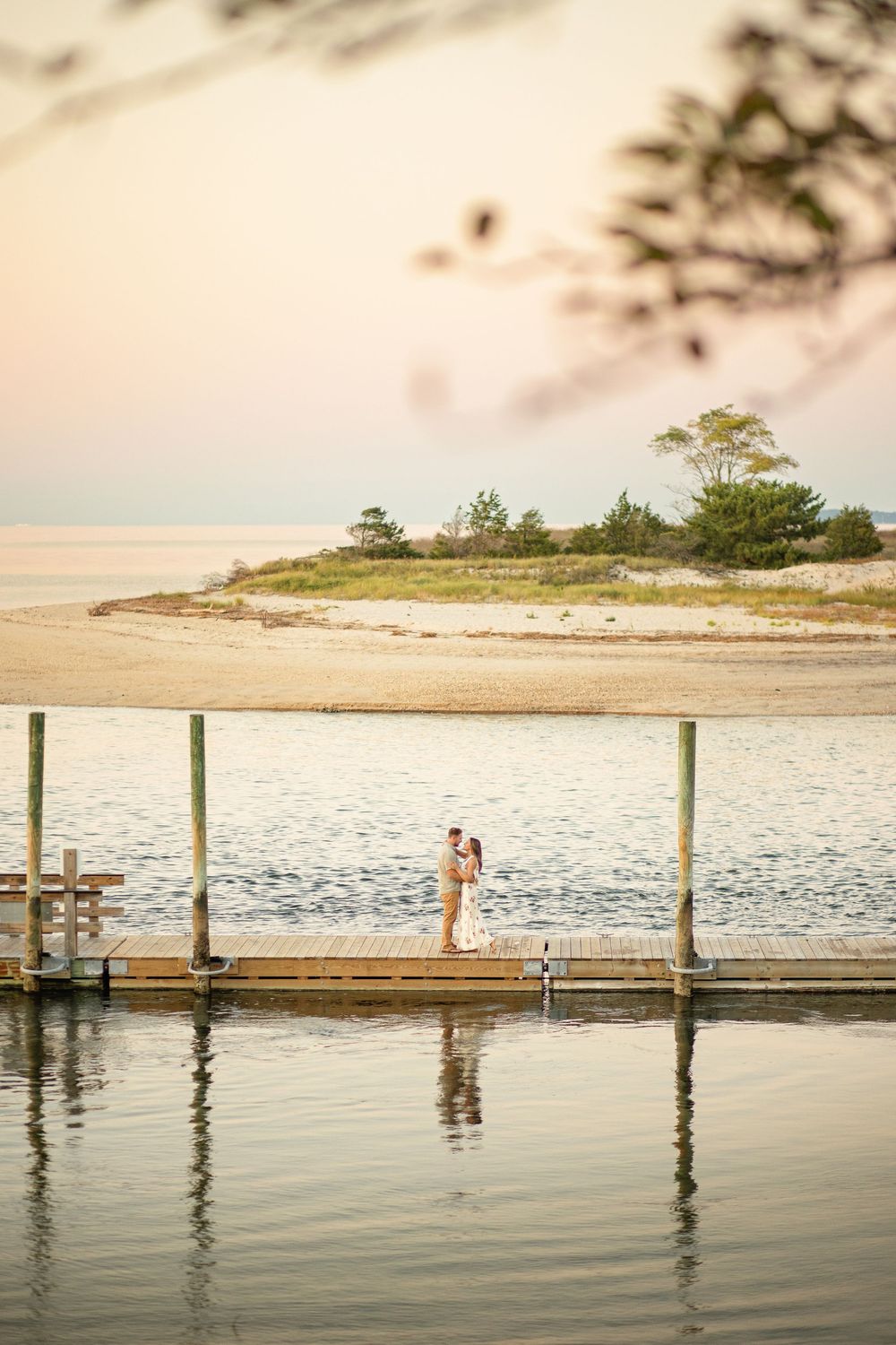 A wooden dock extends over calm waters at sunset with a sandy beach and trees in the background.