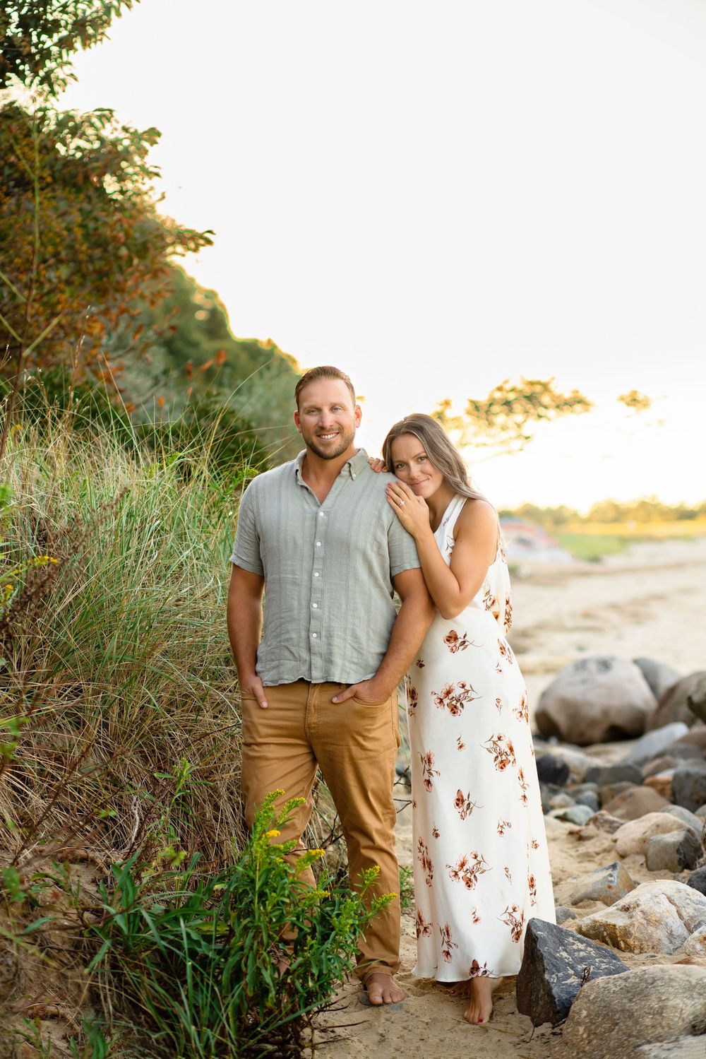 Romantic sunset engagement photos captured on a rocky beach with tall grass and natural coastal scenery.