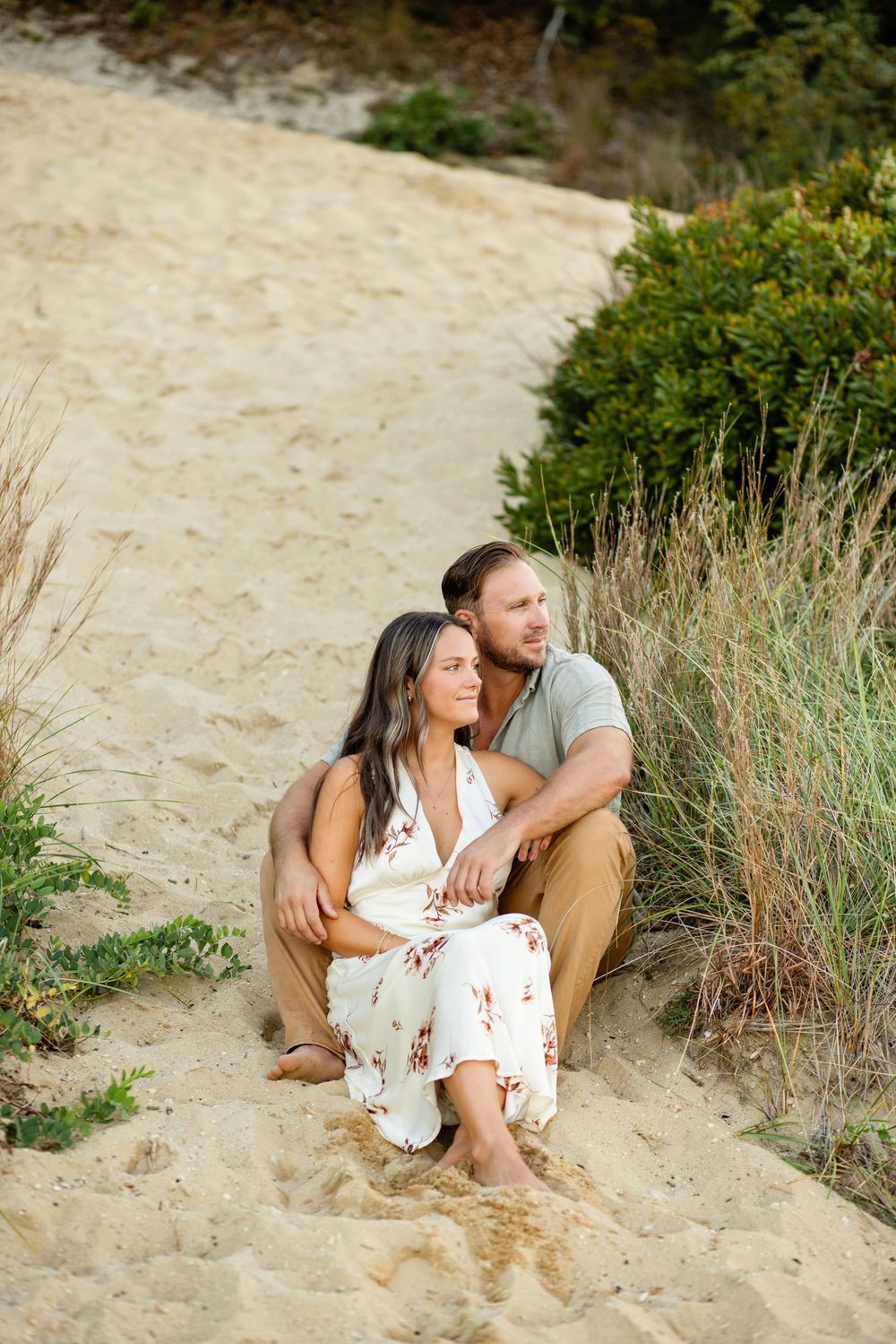 Couple sharing a romantic moment while sitting on a sandy beach slope surrounded by coastal vegetation.