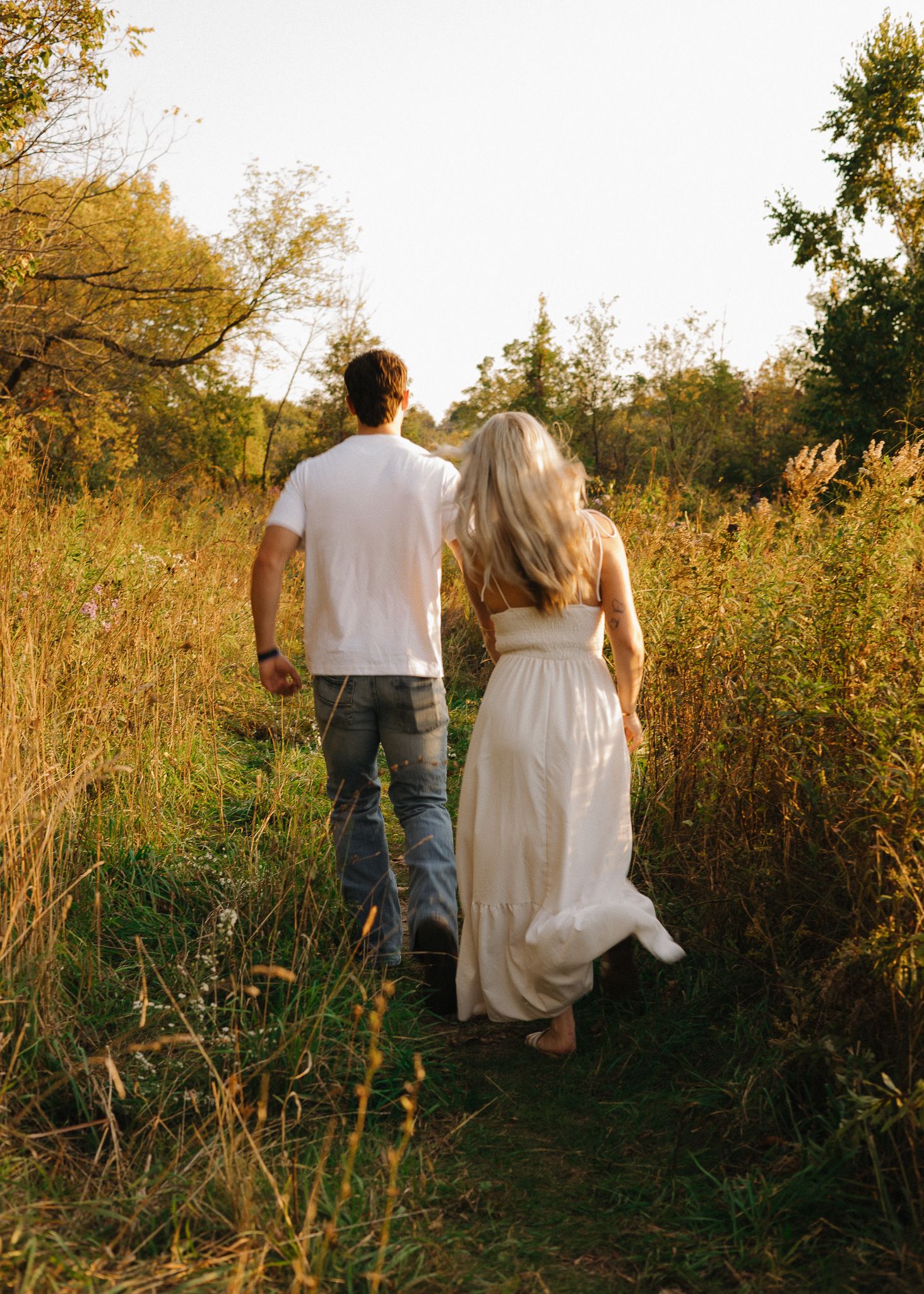 Couple walking hand in hand through a golden meadow path surrounded by tall grasses at sunset.
