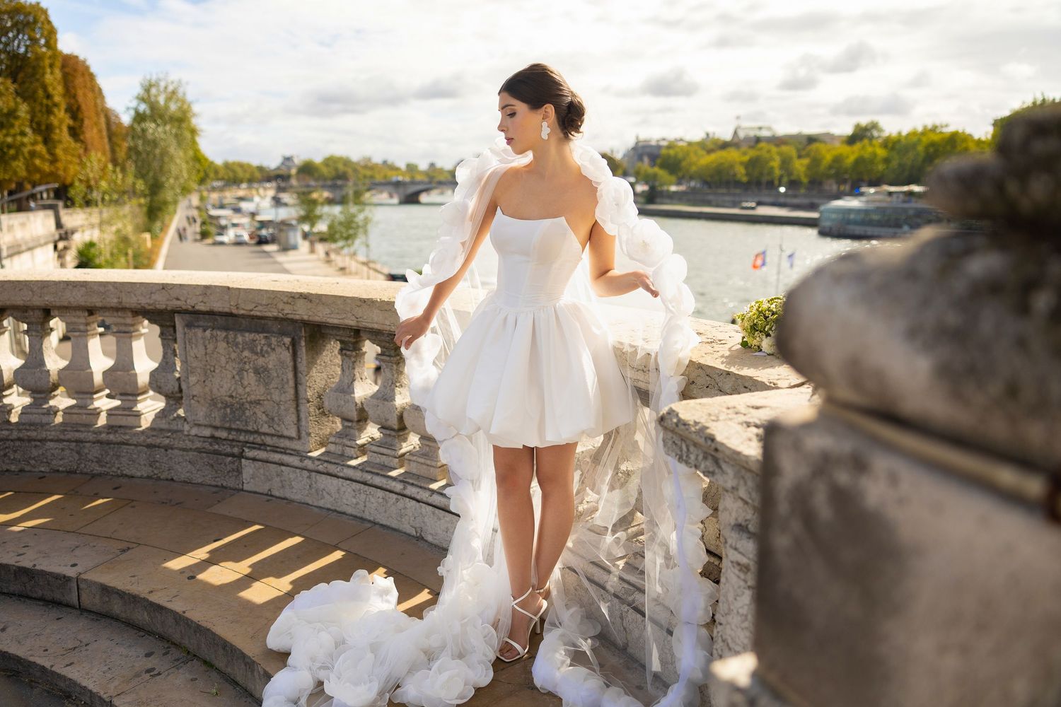 Bride in elegant white dress poses on stone balcony overlooking scenic view.