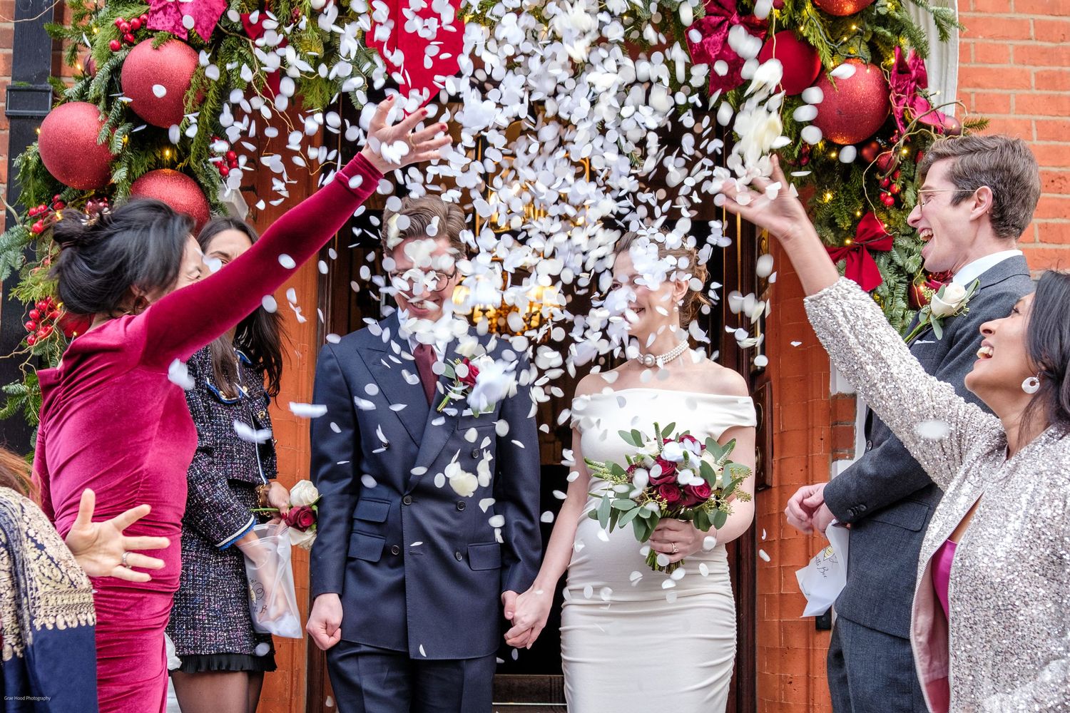 Wedding guests throw white confetti in celebration outside a brick building with pink floral decorations.