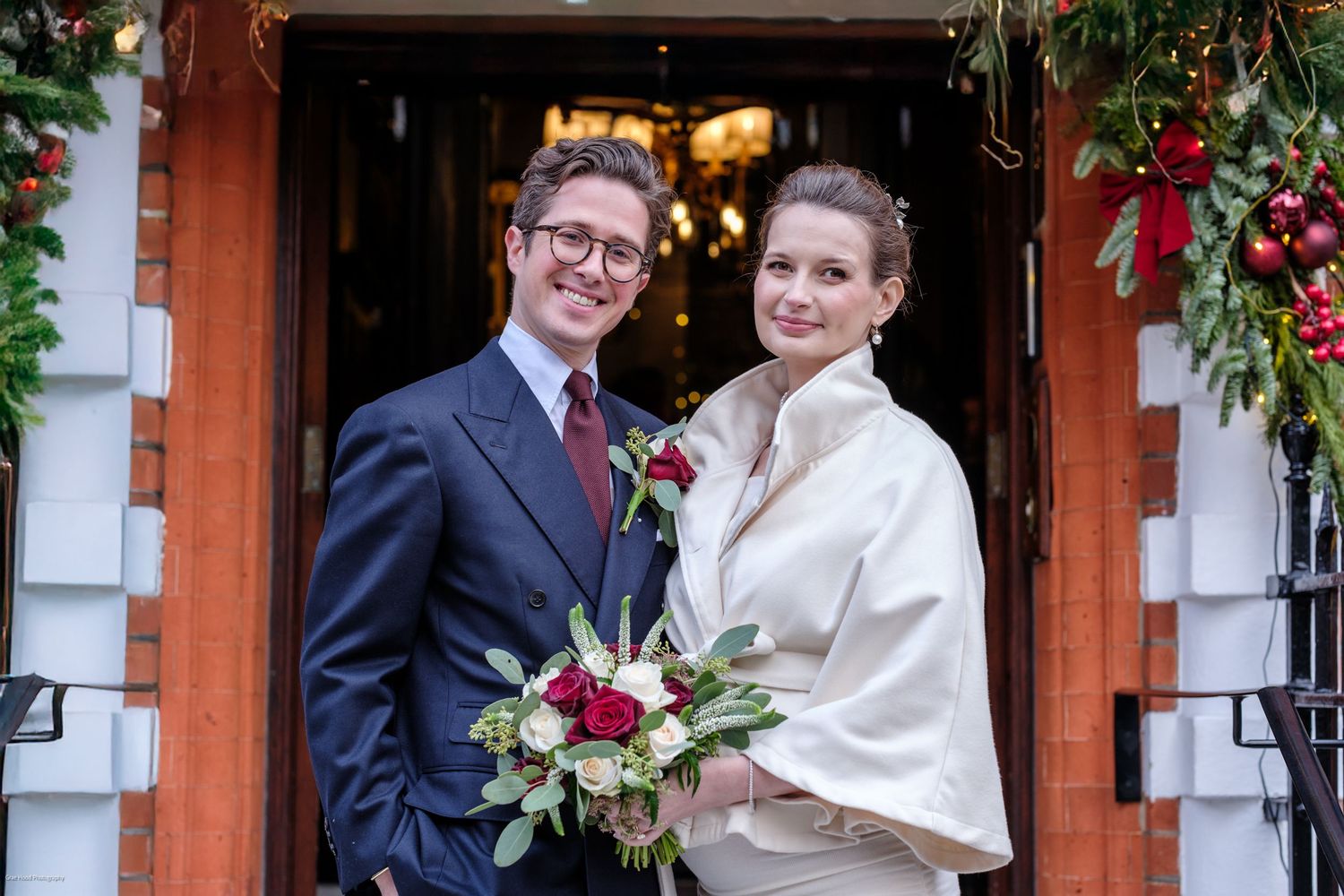 A couple poses for wedding photos in front of a red brick building decorated with festive holiday greenery.