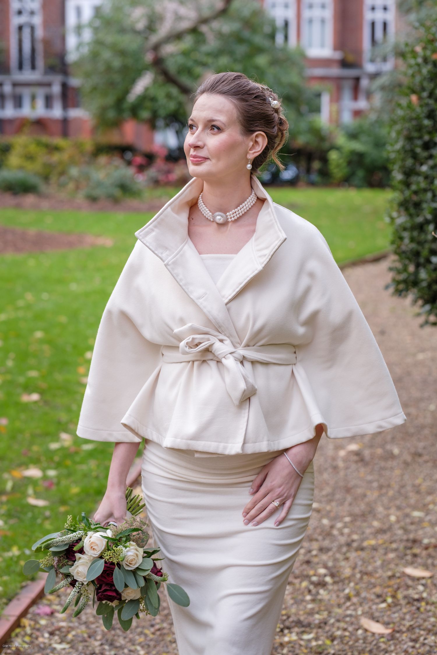Bride in elegant white wrap dress and capelet poses outdoors on garden path with greenery.