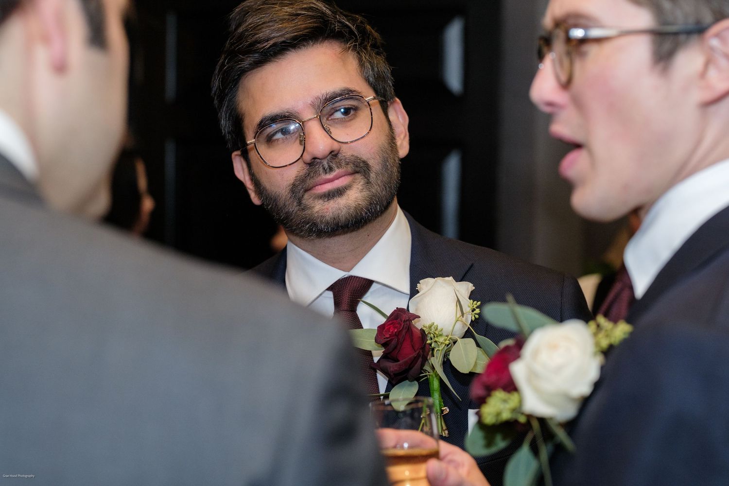 Wedding guests in formal attire pin white rose boutonnieres to their suit lapels during an elegant celebration.
