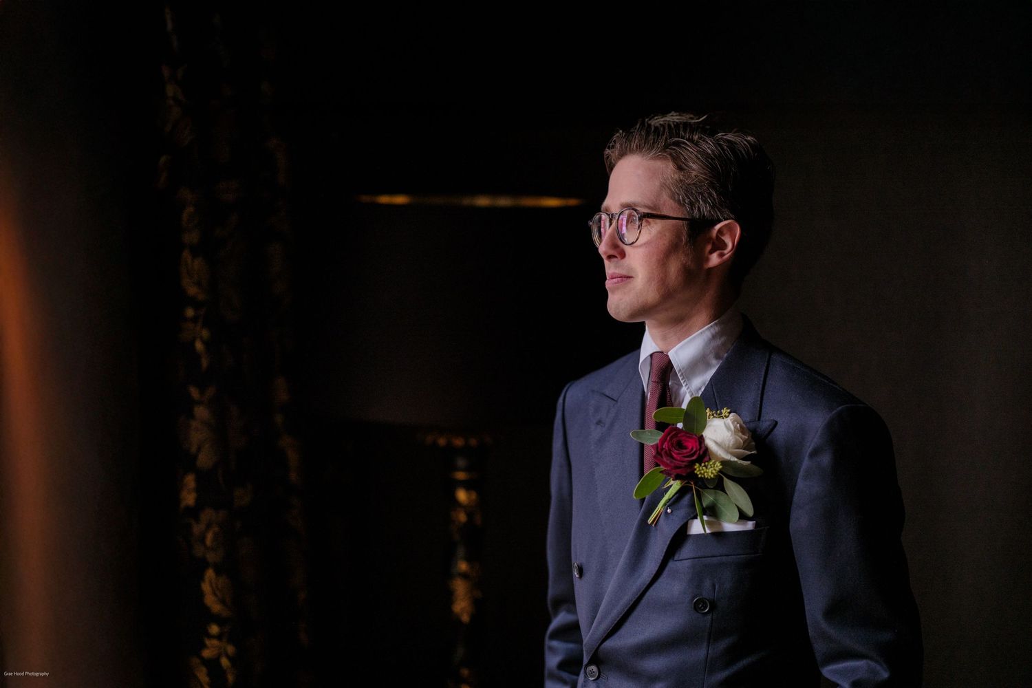 A groom in a navy suit and glasses stands in dramatic lighting with a boutonniere pinned to his lapel.