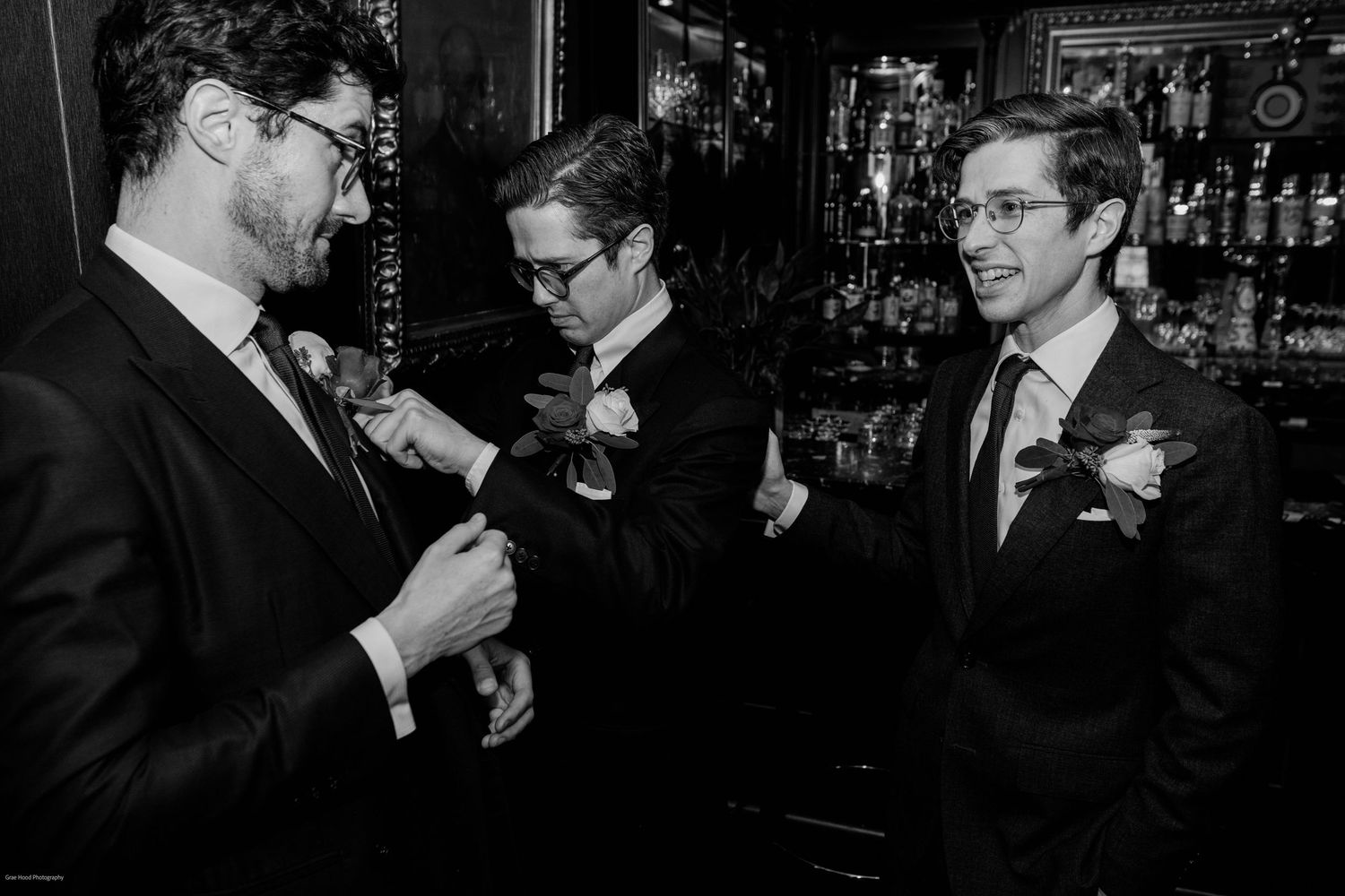 A group of people in formal attire stand at a bar counter having drinks and conversation in a dimly lit setting.